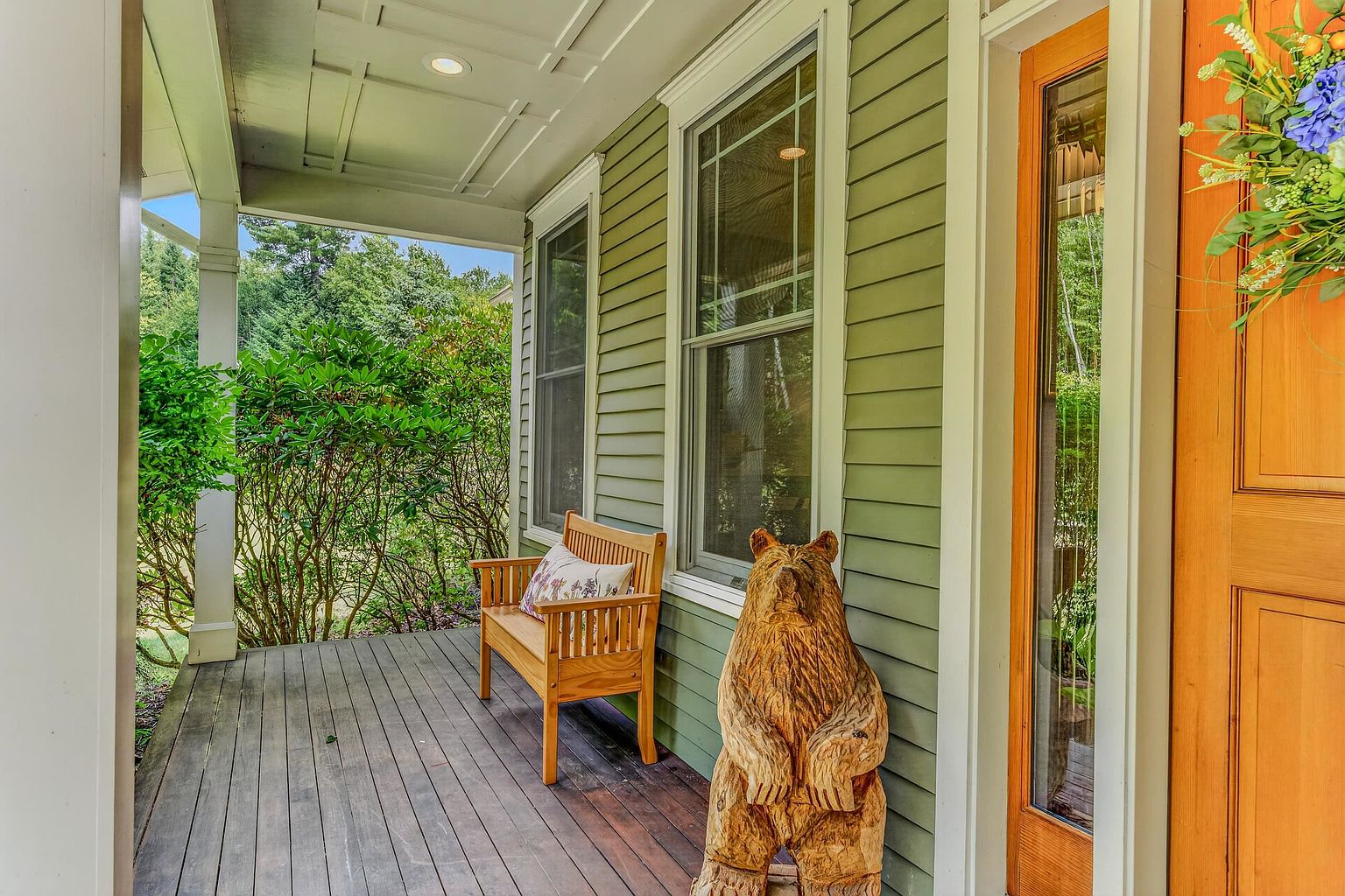 This inviting covered front porch features dark wood decking and light green horizontal siding, creating a warm and rustic aesthetic. A wooden bench with a patterned pillow sits beside a carved wooden bear statue, while a natural wood door with a decorative floral wreath marks the entrance. The perspective looks down the length of the porch, highlighting the architectural details of the ceiling and the lush greenery visible beyond the railing.