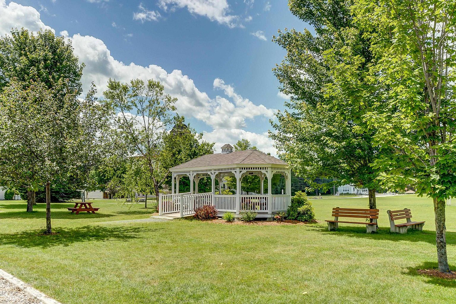 This image captures a serene community park featuring a white, octagonal gazebo as the central focal point, complete with a ramp for accessibility. The well-manicured green lawn is dotted with mature trees, a wooden picnic table, and a park bench, creating a peaceful outdoor gathering space. The perspective is a wide, eye-level shot that emphasizes the open, park-like setting under a bright, partly cloudy sky.