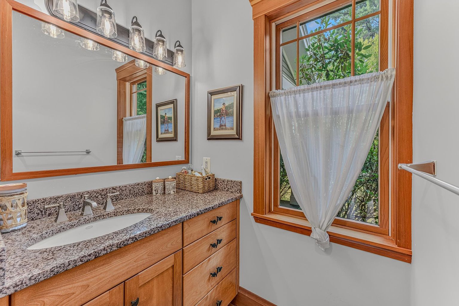 This bathroom features a light wood vanity topped with a speckled stone countertop and an integrated sink. A large mirror reflects the space, which is illuminated by a multi-light fixture, while a window with a white curtain provides natural light. The room is decorated with framed artwork, creating a warm and inviting atmosphere.