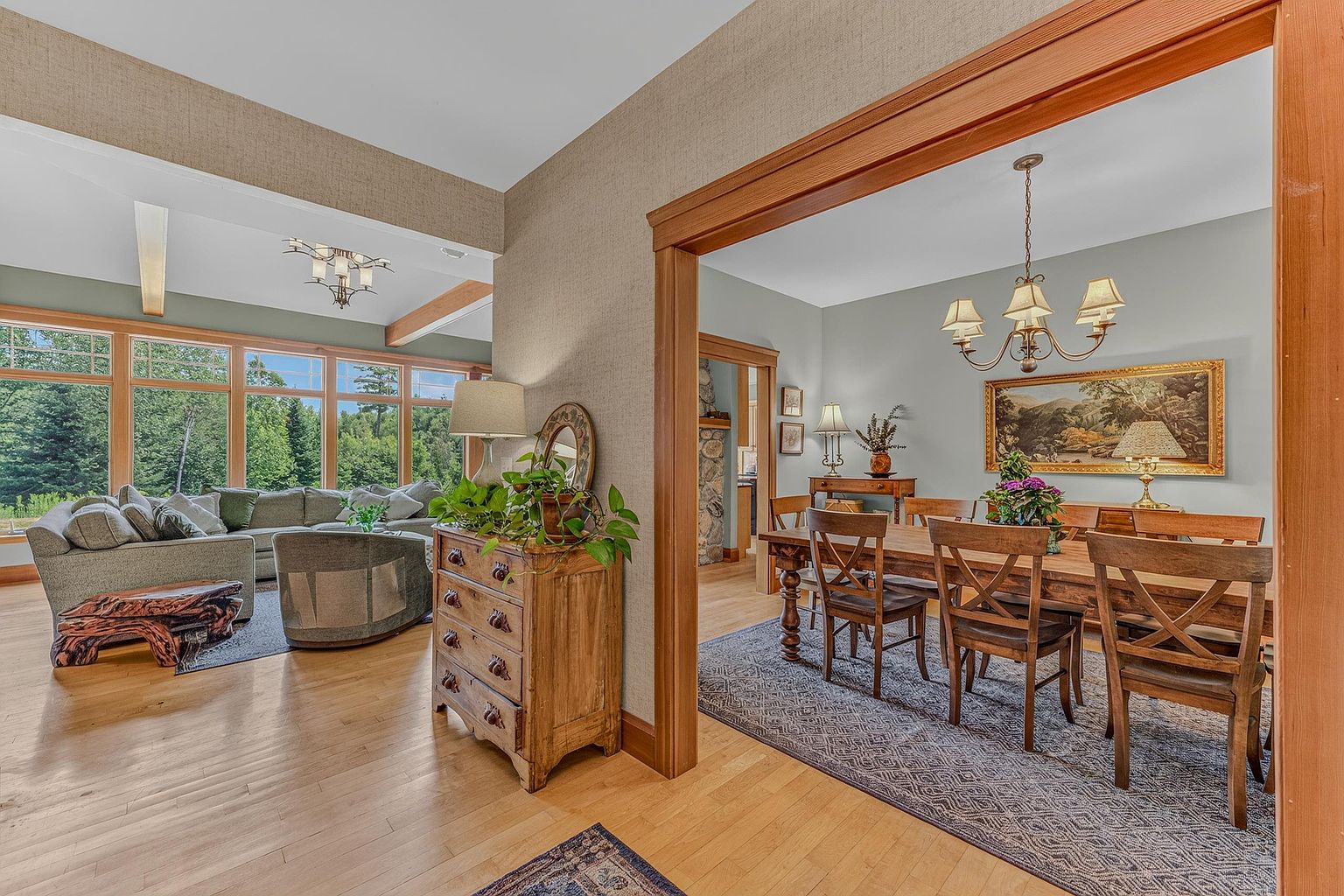 This open-concept living space features light wood flooring and a warm, inviting atmosphere that flows into an adjacent dining area. The living room is anchored by a large, comfortable sectional sofa and a unique, natural-edge wood coffee table, while a rustic wooden chest topped with a lush green plant serves as a focal point. The dining room, framed by a wide wooden archway, showcases a substantial wooden table with matching chairs and a classic chandelier, creating a cohesive and elegant aesthetic throughout the home.