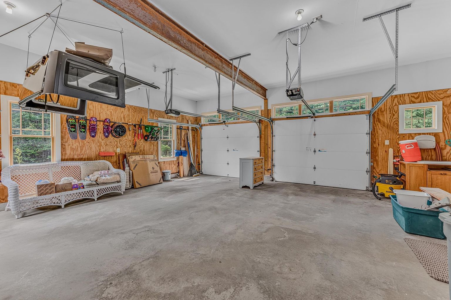 This spacious two-car garage features exposed wood-paneled walls and a concrete floor, providing a rustic and functional workspace. The area includes two white overhead garage doors, a white wicker sofa, and various storage items, including a hardtop suspended from the ceiling. The perspective is wide-angle, capturing the depth and utility of the garage space.