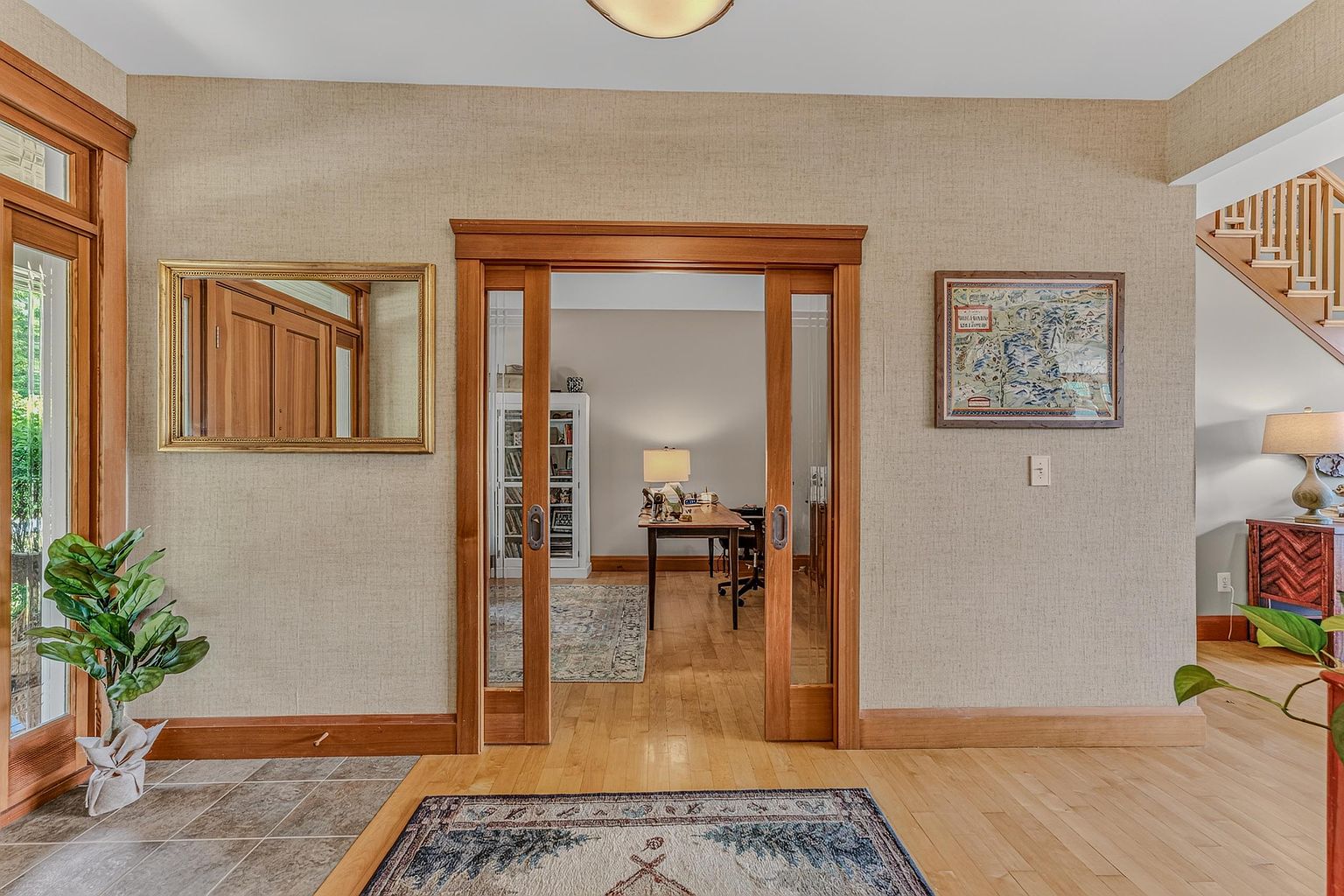This entryway features a warm, inviting atmosphere with light wood flooring and textured neutral wall coverings. A central set of sliding glass doors with wood trim opens into a home office, while a framed map hangs on the wall to the right and a gold-framed mirror sits to the left. The perspective is centered, looking through the doorway toward the office, with a glimpse of a staircase visible on the far right.