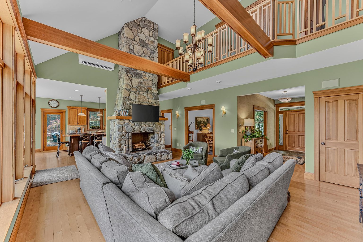 This spacious living room features a prominent stone-clad chimney breast that serves as a central focal point, complemented by a large gray sectional sofa and light wood flooring. The open-concept layout connects to a kitchen area in the background, while a wooden mezzanine balcony overlooks the space from above. The room is bathed in natural light, highlighting the warm wood trim and green wall tones that create a cozy, rustic atmosphere.