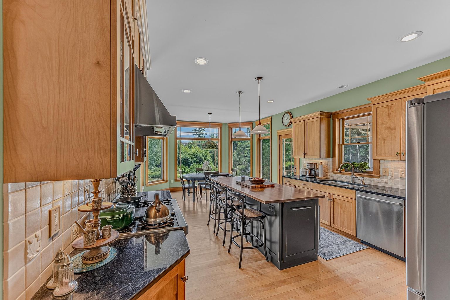 This spacious kitchen features a large central island with a dark base and a wood countertop, complemented by light wood cabinetry and dark stone countertops. The room is bathed in natural light from a series of large windows that offer a scenic view of the surrounding greenery. A stainless steel range hood and appliances add a modern touch to the warm, inviting atmosphere of this well-appointed culinary space.