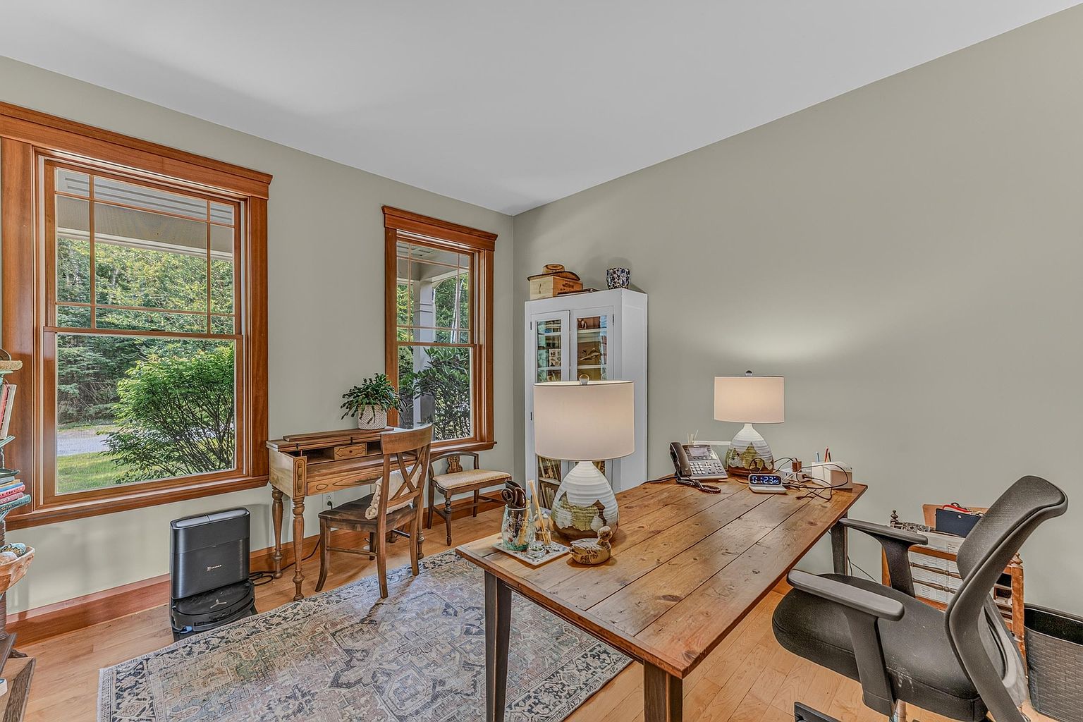 This home office features a rustic wooden desk as the central focal point, complemented by a comfortable ergonomic chair and a secondary writing desk positioned near the windows. The room is bathed in natural light from large windows that overlook the surrounding greenery, creating a serene and productive atmosphere. A patterned area rug anchors the space, while a white cabinet provides storage, contributing to the room's organized and inviting aesthetic.