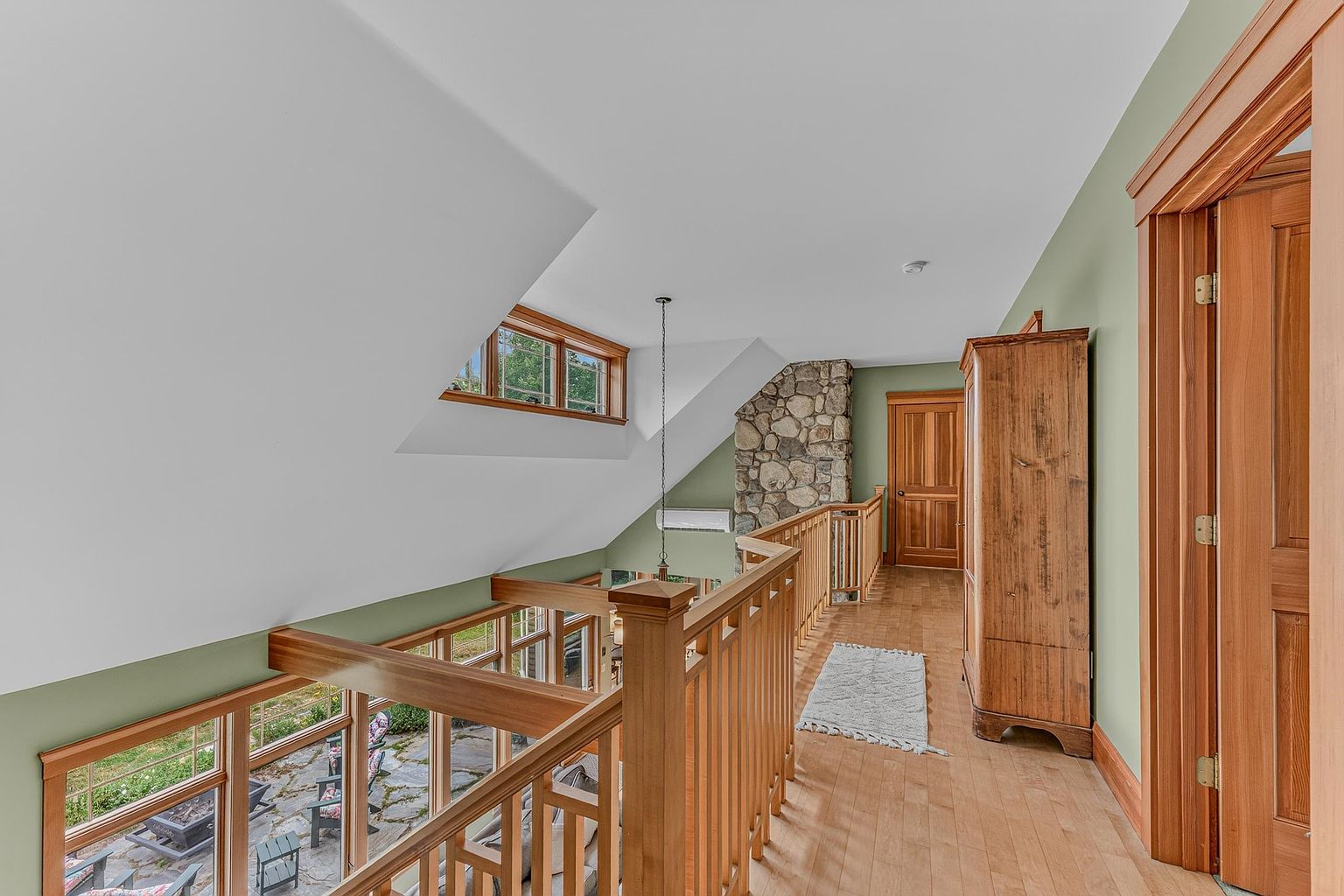 This interior view captures a bright, open-concept hallway overlooking a lower level through a wooden railing. The space features light wood flooring, soft green walls, and a prominent stone-clad chimney breast that adds a rustic, textured element to the design. Large windows allow natural light to flood the area, creating an airy and inviting atmosphere that connects the upper level to the space below.