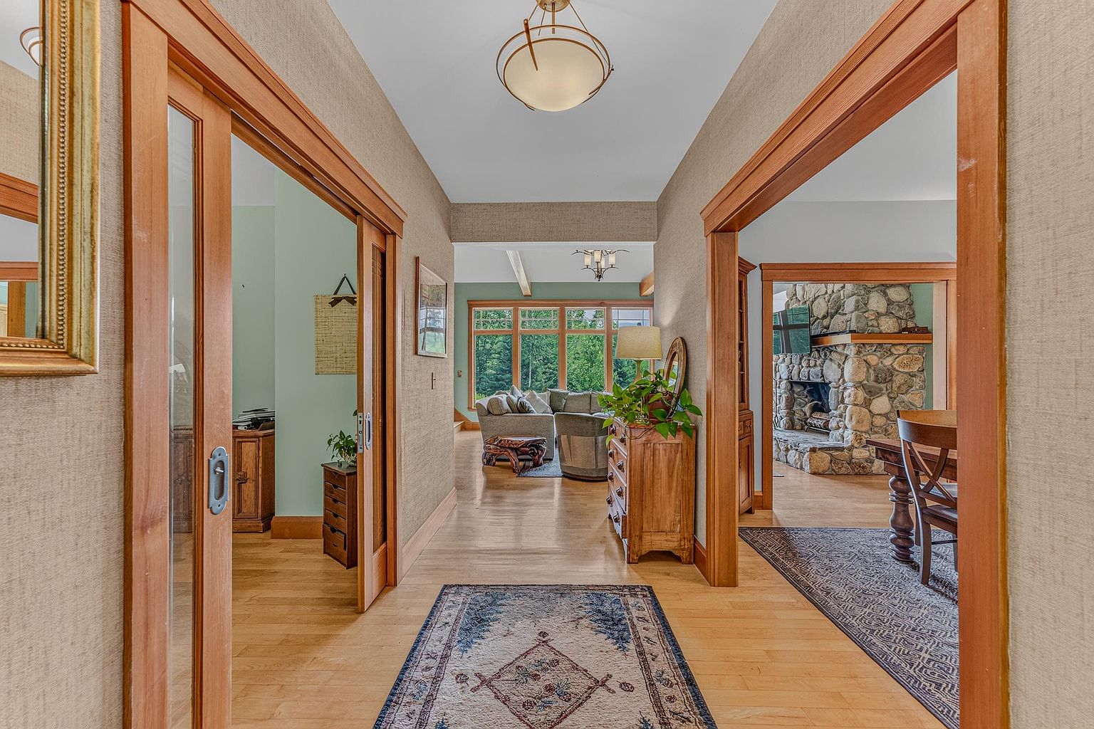 This perspective captures a central hallway that serves as a transition space between multiple rooms, featuring light wood flooring and textured wall coverings. To the left, sliding glass doors open into an office, while to the right, an arched opening leads into a dining area with a prominent stone-clad chimney breast. The hallway is illuminated by a circular flush-mount light fixture and leads the eye toward a bright living area with large windows overlooking the outdoors.