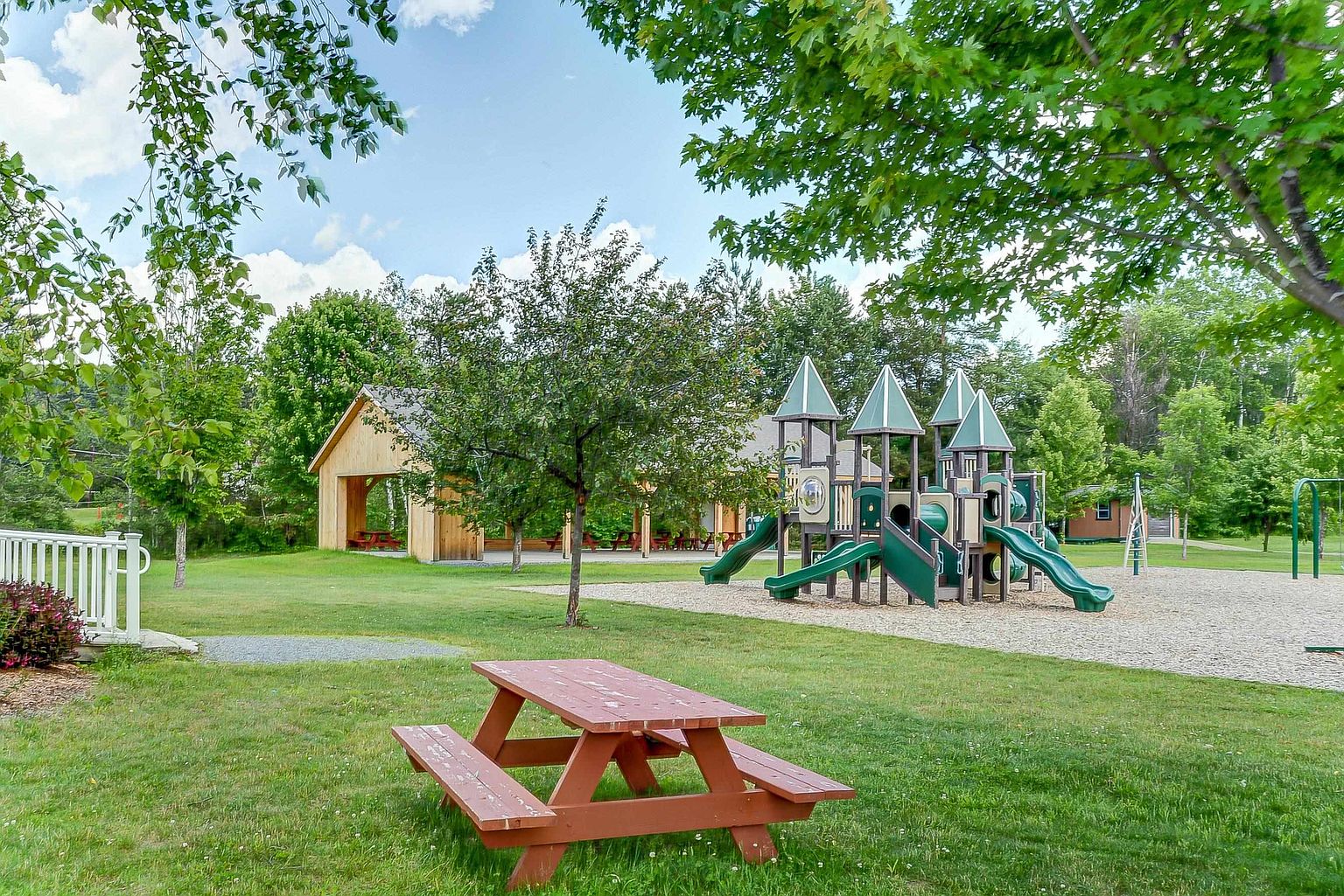 This community park features a well-maintained playground with multiple slides and climbing structures set on a wood-chip base. A wooden pavilion with picnic tables provides a shaded area for gatherings, while a foreground picnic table offers a spot for relaxation on the lawn. The scene is framed by mature trees under a bright, partly cloudy sky, creating an inviting and family-friendly atmosphere.