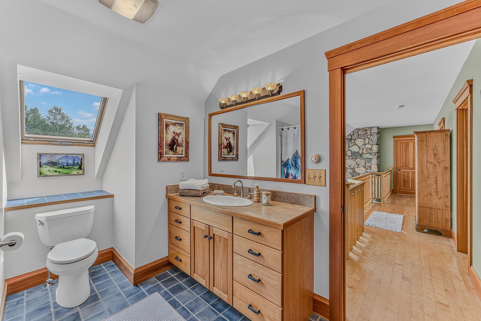 This bathroom features a light wood vanity with a stone countertop and a large mirror illuminated by a multi-bulb light fixture. A toilet is positioned beneath a skylight, and the room is finished with blue tile flooring. The perspective looks into the bathroom from a hallway, revealing a glimpse of a stone-clad wall and wooden flooring in the adjacent space.