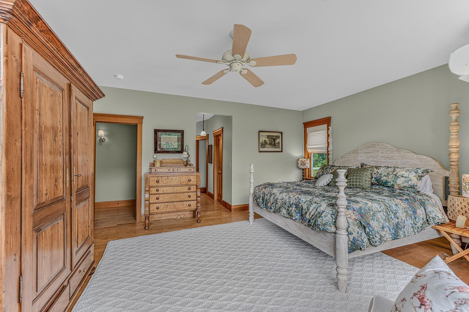 This spacious primary bedroom features soft sage green walls, light wood flooring, and a large, textured area rug. The room is anchored by a substantial light-toned wooden bed frame with a patterned floral duvet, complemented by a large, rustic wooden armoire and a matching dresser. The perspective captures the room from a slightly elevated angle, showcasing the warm, traditional aesthetic and the transition into the adjacent hallway.