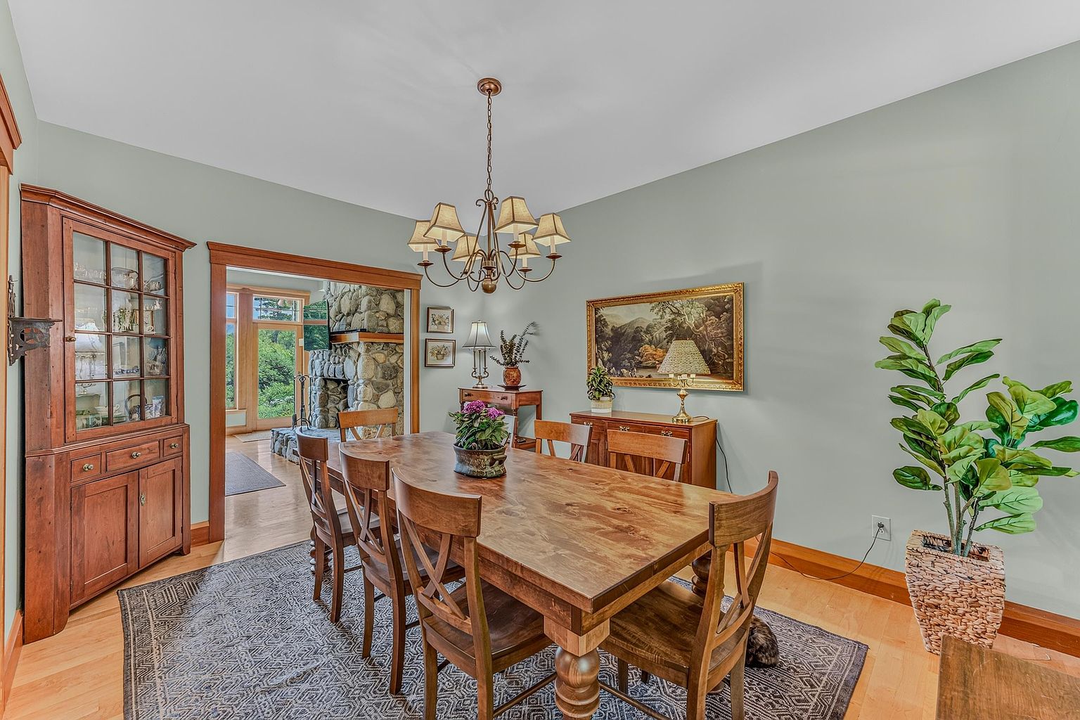 This formal dining room features a warm, traditional aesthetic with light green walls and natural wood trim. A large wooden dining table serves as the centerpiece, illuminated by a multi-armed chandelier, while a matching hutch and sideboard provide ample storage and display space. Through the doorway, a glimpse of a stone-clad chimney breast in the adjacent room adds a rustic architectural element to the home's open flow.