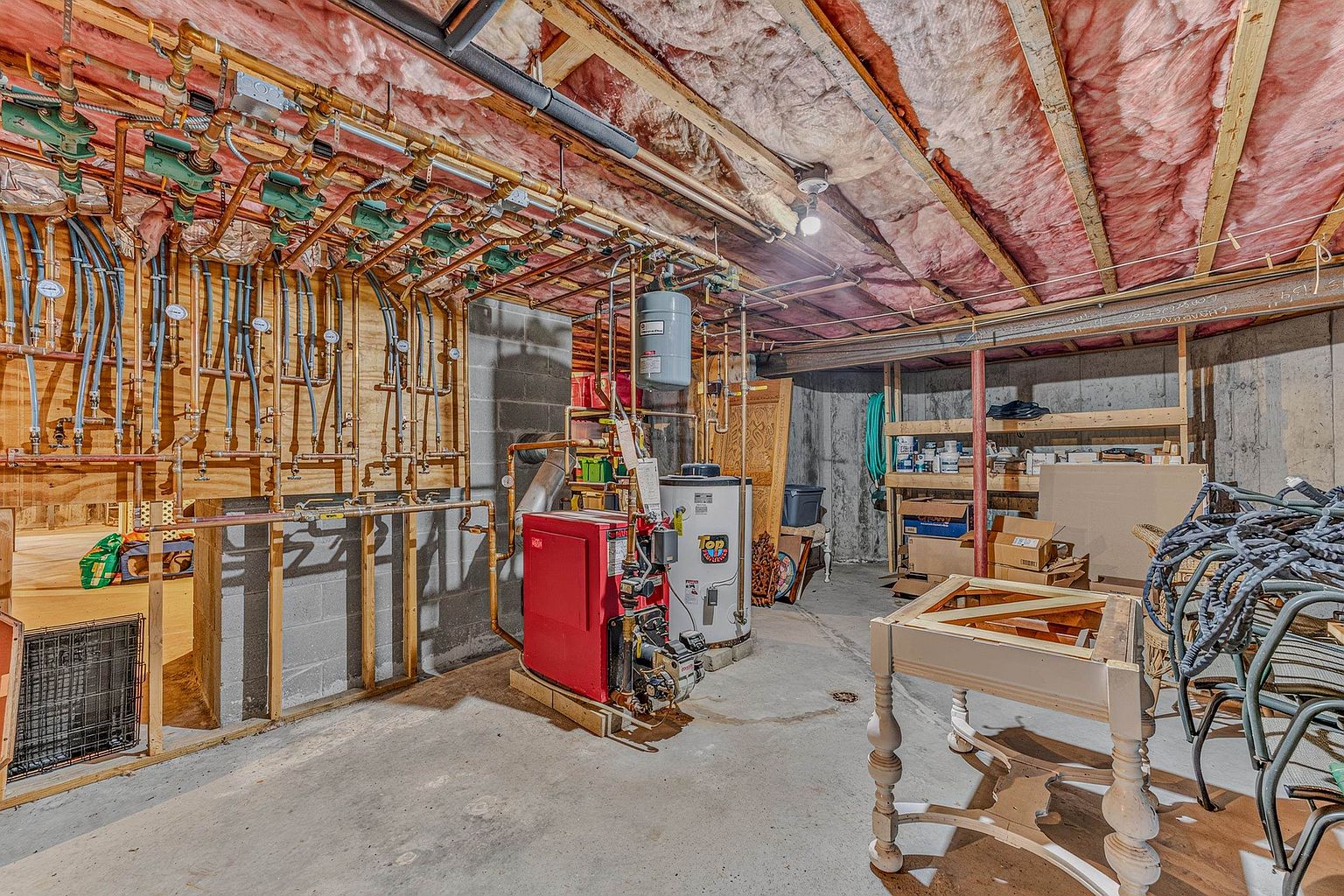 This unfinished basement utility area features exposed ceiling joists with pink insulation and a concrete floor. The space is dominated by a complex hydronic heating system with numerous copper pipes, valves, and pumps mounted on a wooden wall panel, alongside a red boiler and a white water heater. To the right, wooden shelving units provide storage, and a partially assembled table sits in the foreground, suggesting a functional, utilitarian space.