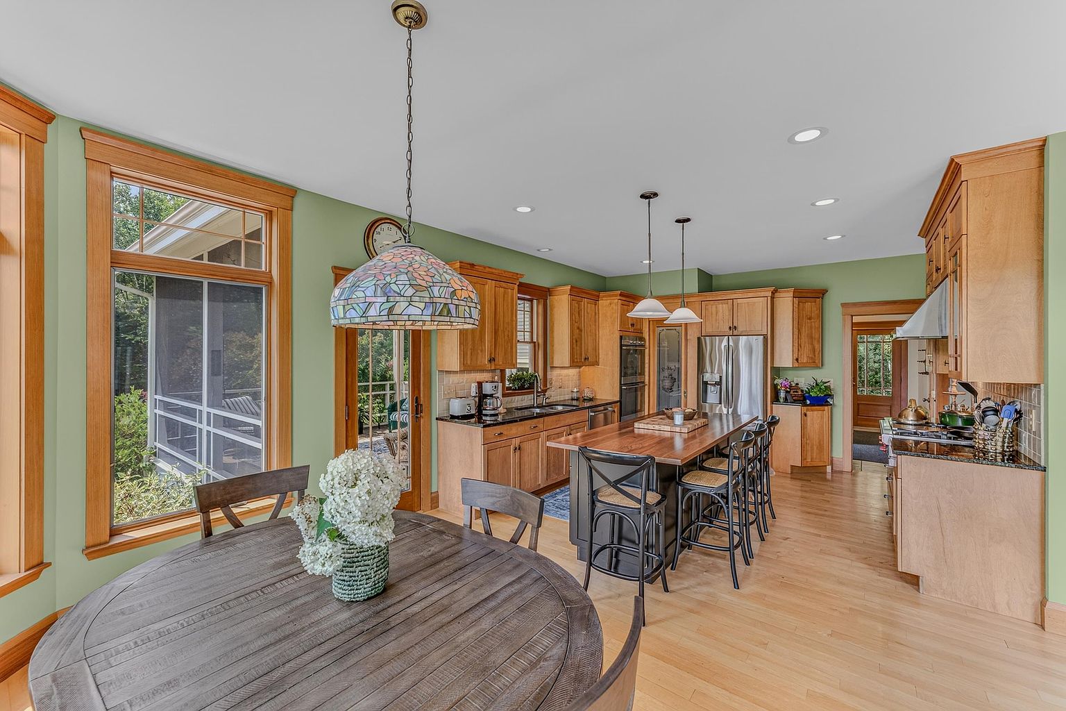 This spacious, open-concept kitchen features warm-toned wood cabinetry, a large central island with a dark base and wood countertop, and light-colored hardwood flooring. A round dining table sits in the foreground under a decorative stained-glass pendant light, while the kitchen area is illuminated by recessed lighting and two additional pendant lights over the island. The space is painted a soft green, creating a welcoming atmosphere that flows seamlessly into the adjacent dining area.