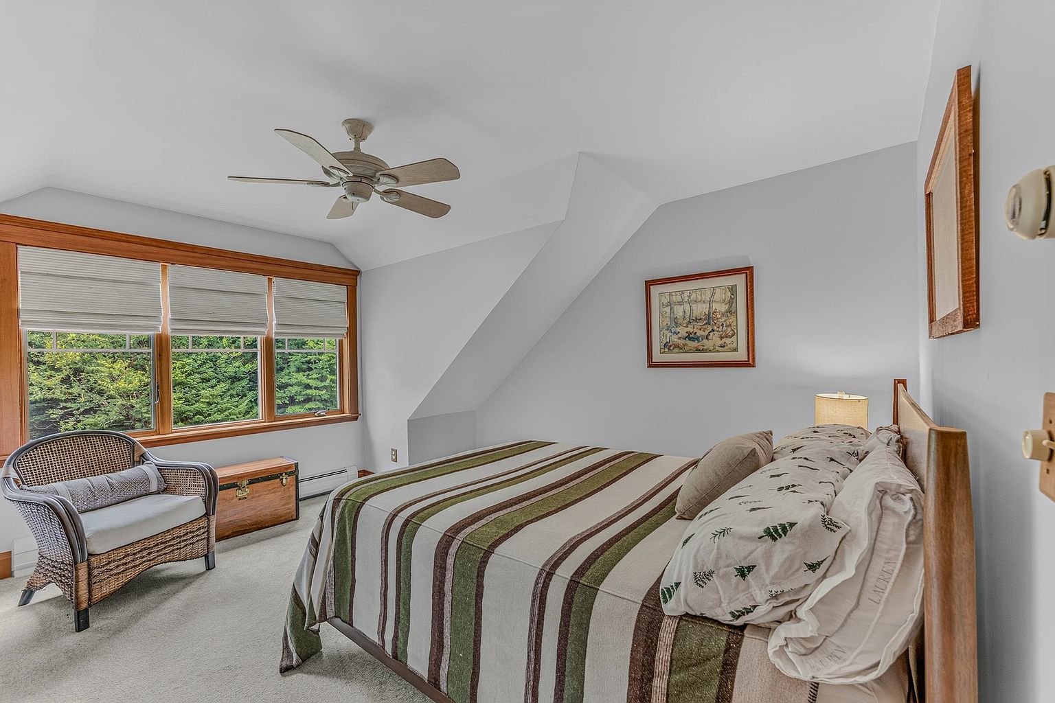This cozy bedroom features a bed with a striped comforter and patterned pillows, positioned against a light-colored wall. A wicker armchair sits near a large window with wooden trim, providing a view of the surrounding greenery, while a wooden chest adds a rustic touch to the space. The room is illuminated by a ceiling fan and a bedside lamp, creating a warm and inviting atmosphere.
