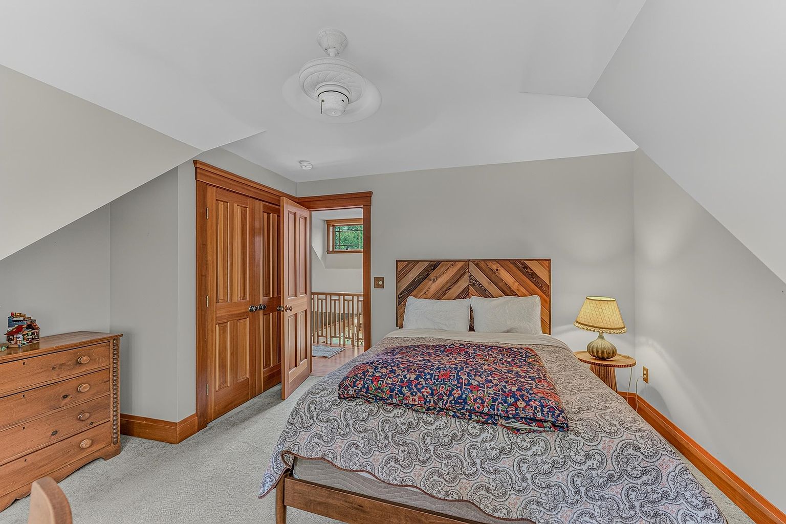 This cozy bedroom features a bed with a distinctive chevron-patterned wood headboard and a colorful patterned quilt. The room is accented by warm wood trim around the doors and baseboards, a wooden dresser, and a small bedside table with a lamp. A doorway leads to an adjacent hallway with a railing, providing a view into the space from a slightly elevated perspective.