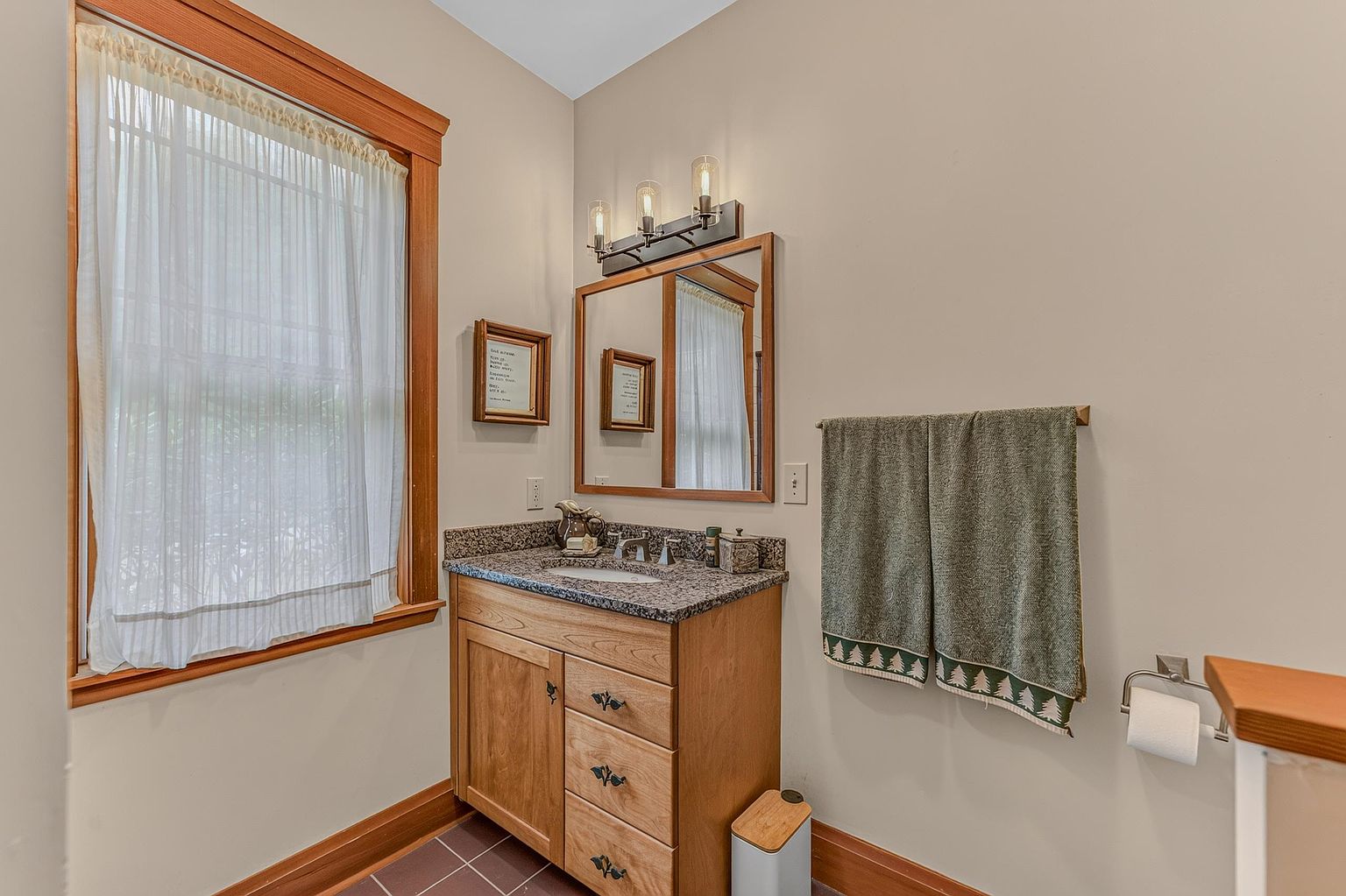 This compact bathroom features a light wood vanity topped with a speckled stone counter and an integrated sink. A rectangular mirror is mounted above the vanity, flanked by a three-light fixture, while a window with sheer curtains provides natural light. The space is finished with a towel rack holding a patterned towel and a wall-mounted toilet paper holder, creating a functional and tidy guest space.