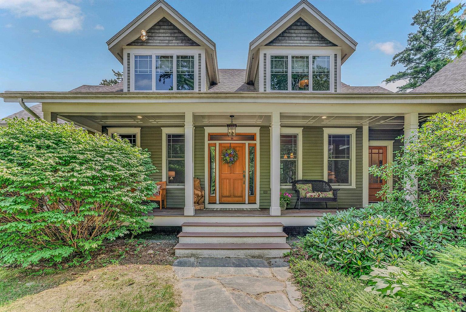 This charming, green-sided home features a welcoming covered front porch with white columns and a natural wood front door adorned with a floral wreath. Two symmetrical dormers with multi-pane windows sit above the porch, adding architectural interest to the roofline. A stone walkway leads up to the wooden steps, flanked by lush green landscaping and mature shrubs that frame the entrance.