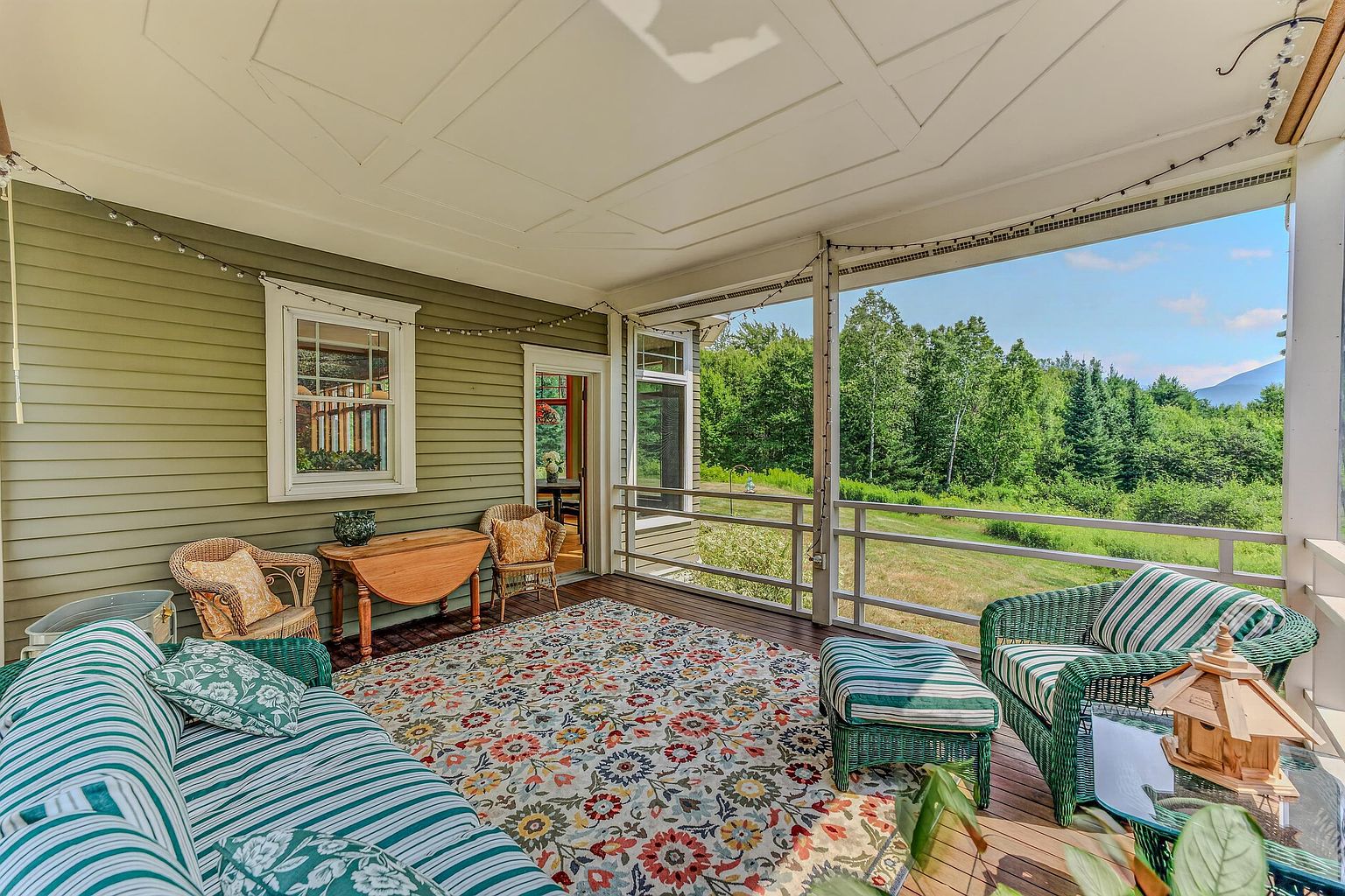 This inviting screened-in porch features a comfortable seating area with green and white striped wicker furniture and a vibrant, multi-colored floral area rug. The space is finished with olive-green siding, a decorative ceiling, and a wooden drop-leaf table, offering a serene view of the surrounding lush forest and meadow. The open-air design creates a seamless transition between the indoor living space and the natural landscape, perfect for relaxing or entertaining.