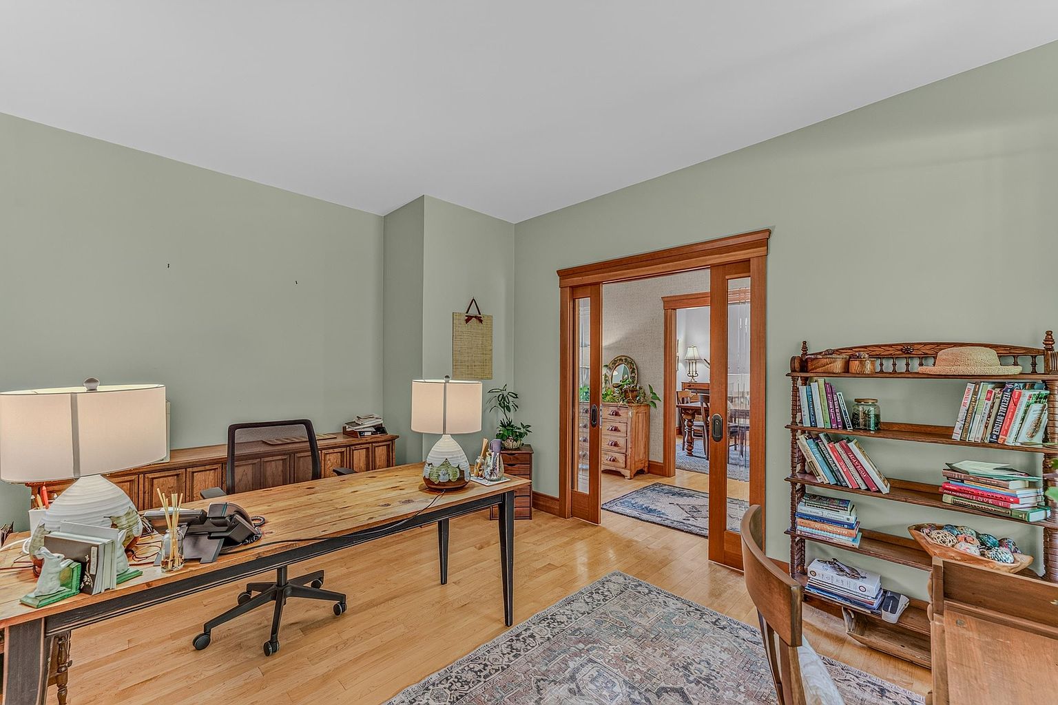 This home office features light green walls, hardwood flooring, and a large wooden desk positioned centrally in the room. A wooden bookshelf filled with books stands to the right, while a sliding wooden door leads into an adjacent dining area. The space is well-lit by two table lamps, creating a warm and productive atmosphere.