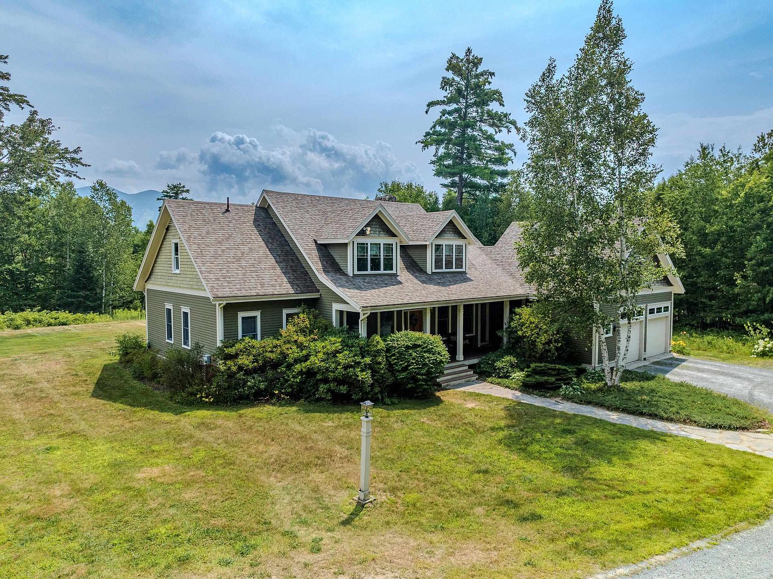 This elevated perspective captures a charming, green-sided home nestled in a lush, wooded landscape. The house features a multi-gabled roofline with dormer windows, a covered front porch, and an attached garage, all set against a backdrop of rolling hills and a clear blue sky. A stone walkway leads to the entrance, surrounded by mature landscaping and a sprawling lawn, creating a serene and inviting curb appeal.