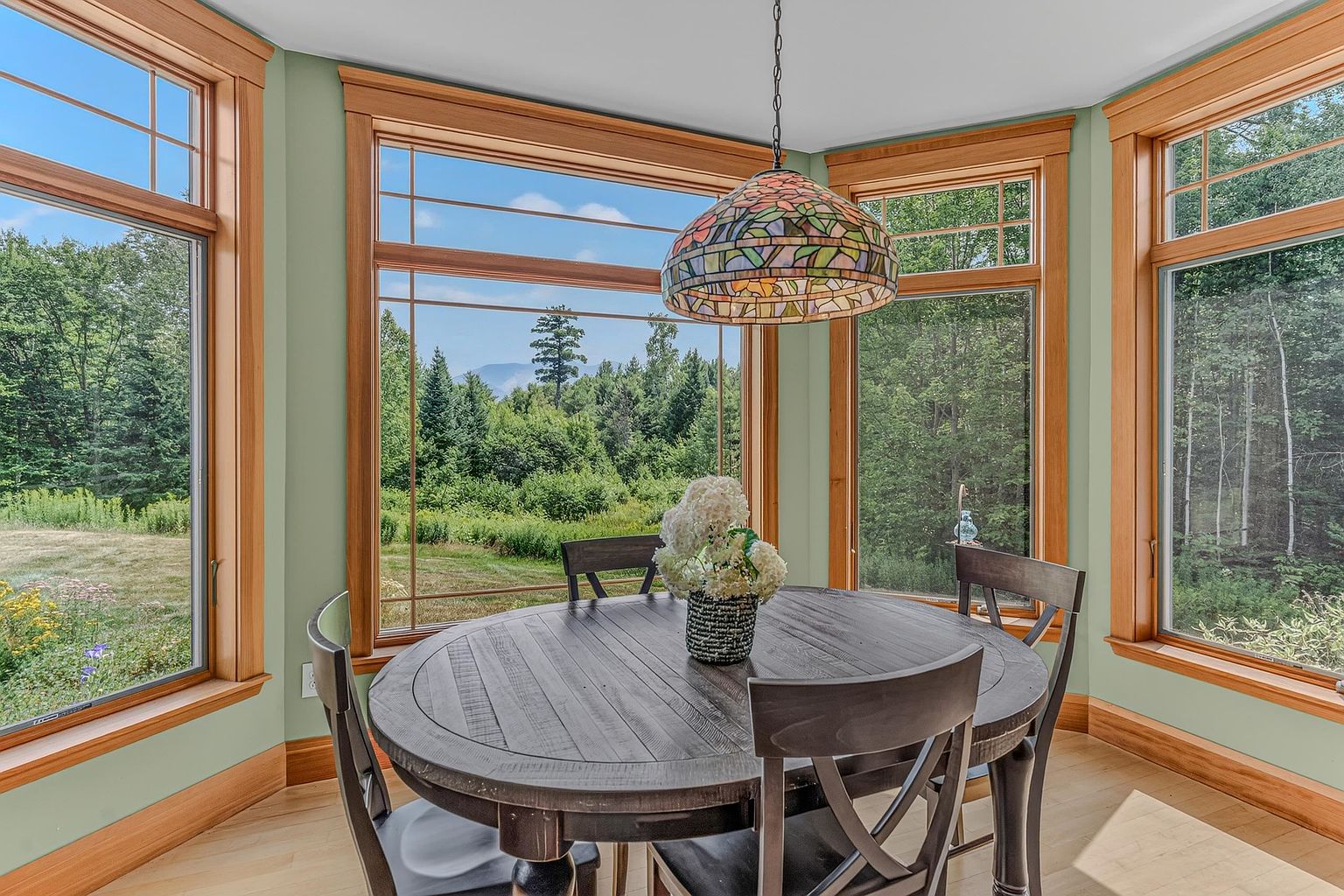 This bright dining area features a round dark wood table surrounded by matching chairs, positioned beneath a decorative stained-glass pendant light. The room is framed by large windows with warm wood trim that offer expansive, scenic views of the surrounding forest and distant mountains. The light green walls and light-toned wood flooring create a serene and inviting atmosphere perfect for casual meals.