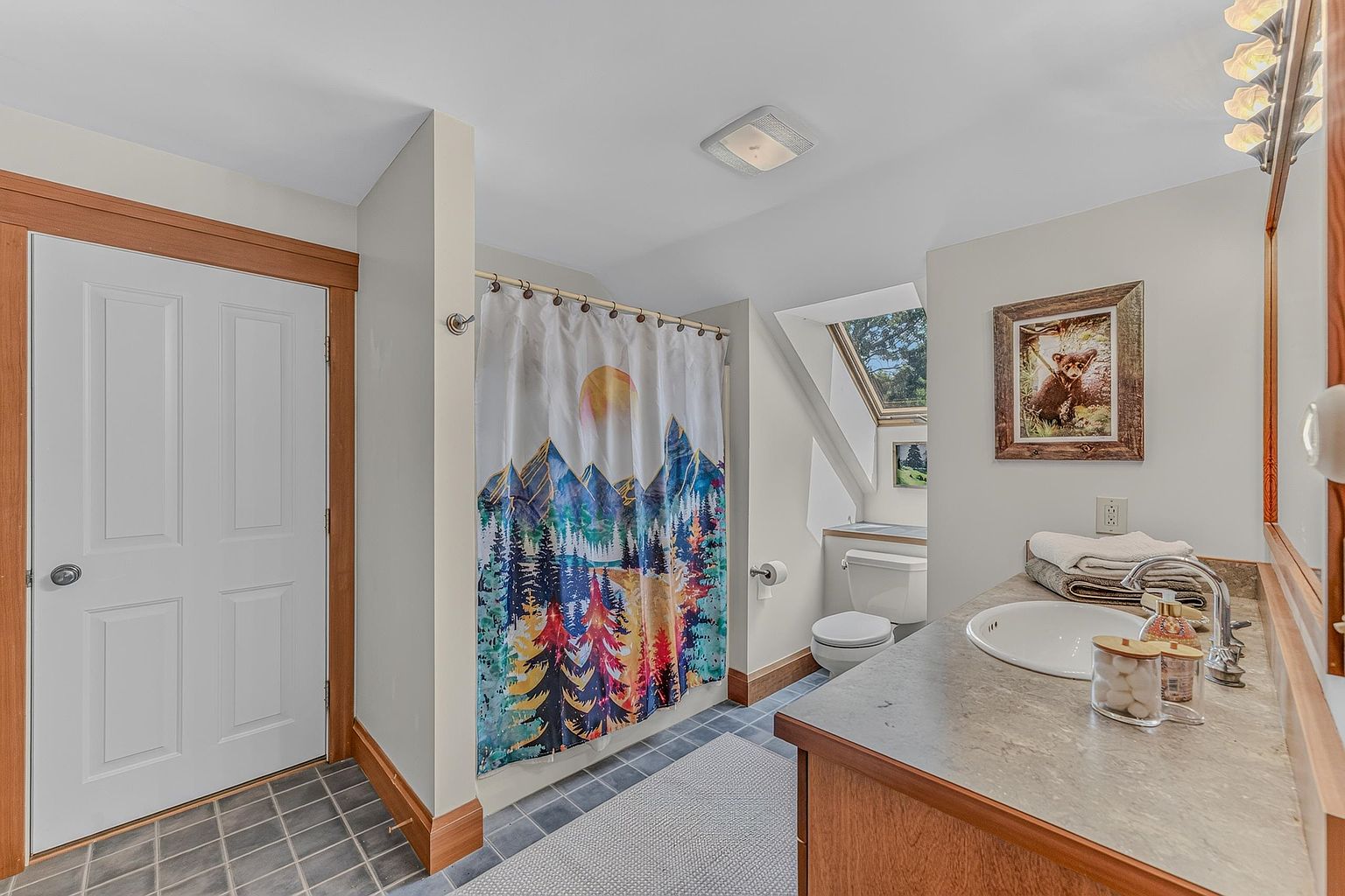 This bathroom features a rustic, nature-inspired aesthetic with a shower curtain depicting a mountain landscape and a framed bear print on the wall. The space includes a vanity with a neutral countertop, a white sink, and a skylight that brings natural light into the toilet area. The room is finished with dark square floor tiles and light-toned walls, creating a cozy and functional atmosphere.
