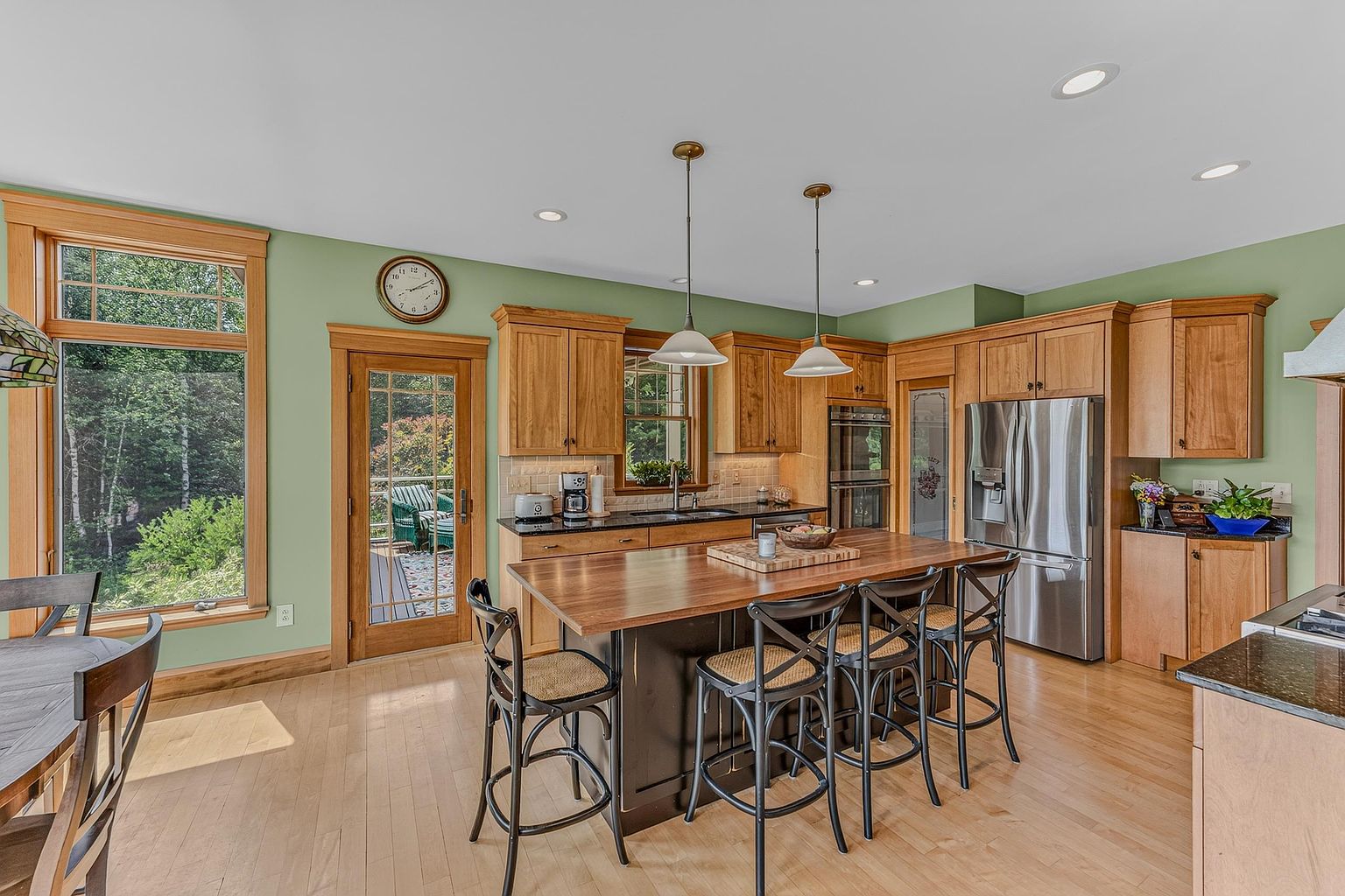 This bright and inviting kitchen features a large central island with a dark base and a warm-toned wood countertop, complemented by matching light wood cabinetry. The space is accented by sage green walls, stainless steel appliances, and two pendant lights hanging over the island. A glass-paneled door and large windows provide a seamless connection to the outdoors, creating an airy and functional atmosphere.
