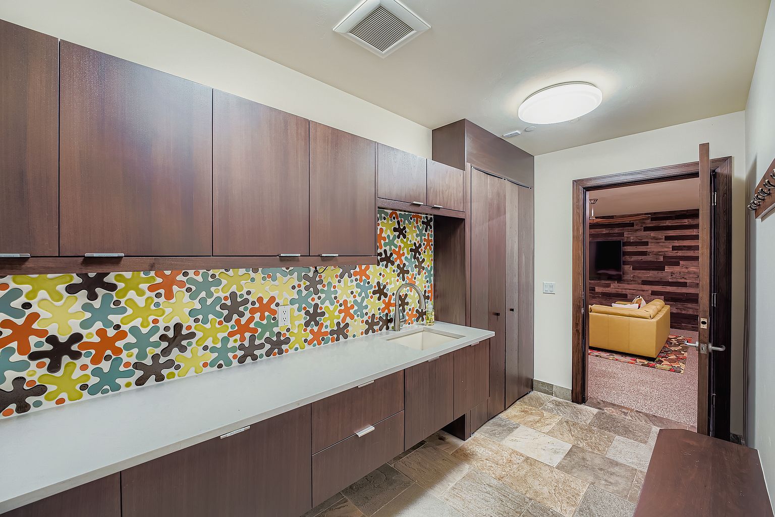 This interior shot showcases a modern laundry room with dark wood cabinetry and a colorful backsplash. A white countertop provides ample workspace above the lower cabinets, and a stainless steel faucet complements the sink. The room features tile flooring and an open doorway leading to another room, suggesting a well-designed and functional space.