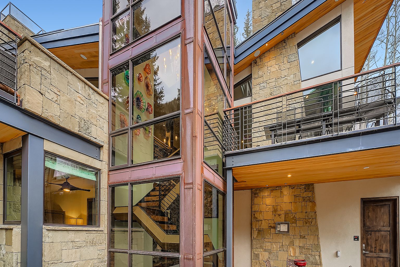 This is a side view of a modern, multi-story home featuring a unique architectural design. The exterior is characterized by a combination of stone cladding, large windows with copper-colored frames, and metal accents. A glass-enclosed staircase is a prominent feature, and a balcony with metal railings adds to the home's appeal.