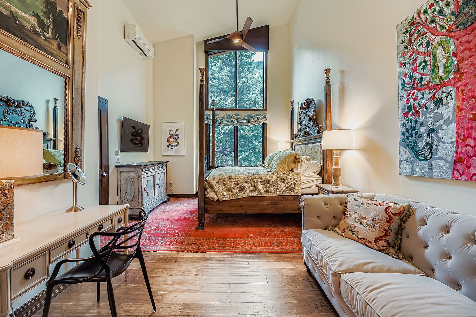 This is a beautifully decorated primary bedroom featuring a four-poster bed with ornate detailing and luxurious bedding. The room is accented with a vibrant red rug, a comfortable sofa, and unique artwork, creating a sophisticated and inviting atmosphere. Natural light floods the space through a large window, enhancing the room's warmth and elegance.