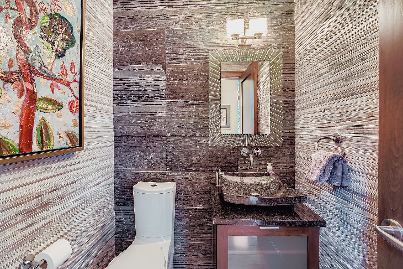 This is a well-appointed guest bathroom featuring a unique vessel sink atop a dark wood vanity. The walls are adorned with horizontally-laid textured tiles, complemented by a decorative mirror and modern lighting fixture. A white toilet sits adjacent to the vanity, and an artistic painting hangs on the wall, adding a touch of elegance to the space.