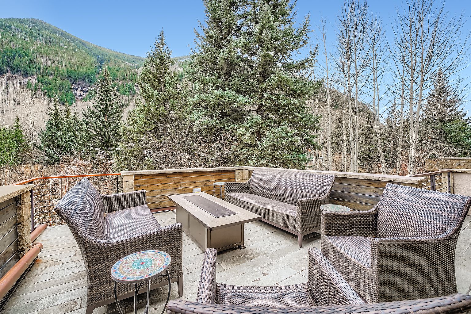 This image showcases a beautifully designed outdoor patio or deck area, furnished with comfortable wicker seating arrangements around a modern fire pit. The space is surrounded by a wooden railing and offers a scenic view of the surrounding mountains and trees, creating a serene and inviting atmosphere. The stone flooring adds a touch of rustic elegance to the outdoor living space.