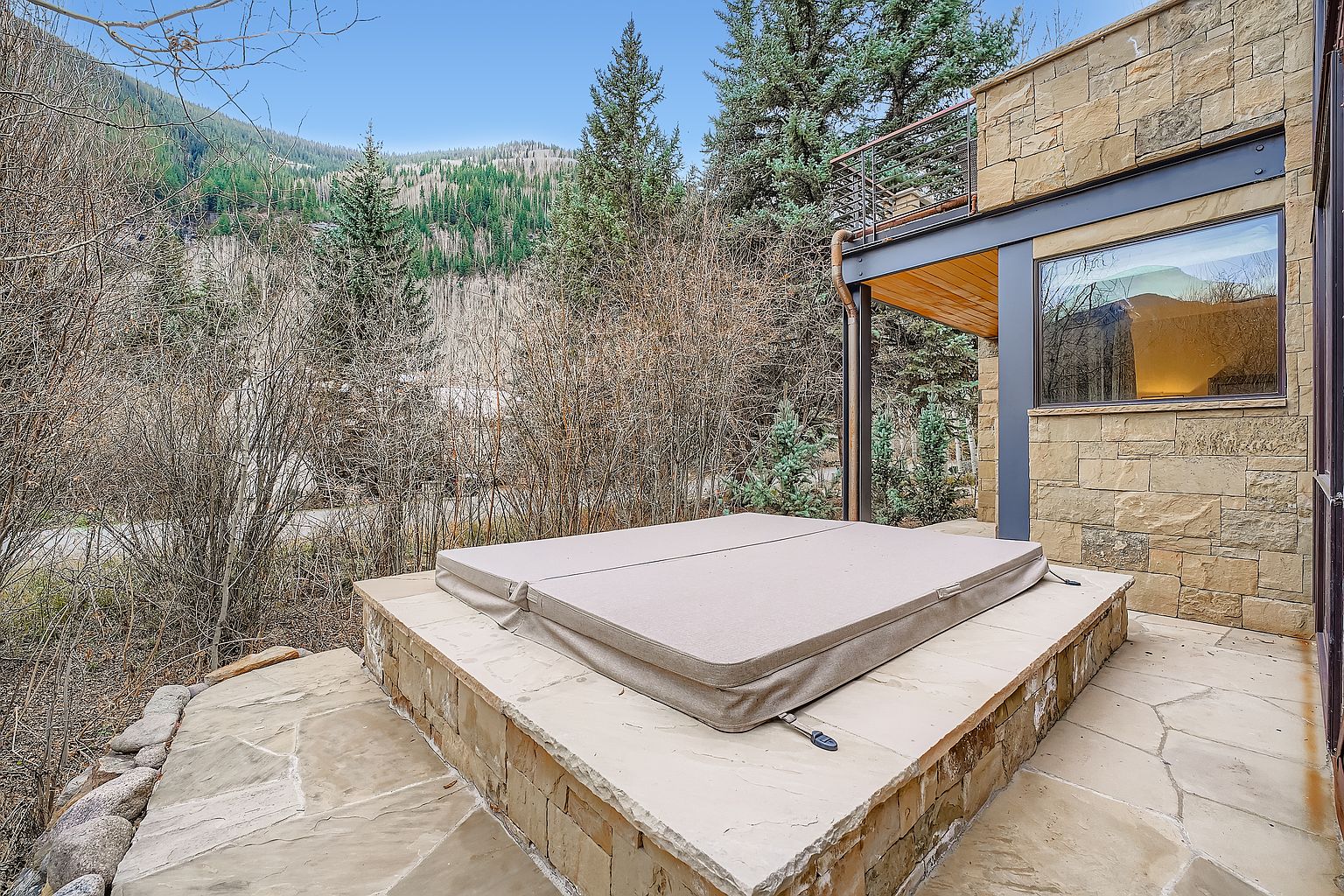 This image showcases an exterior patio area featuring a built-in hot tub with a beige cover. The patio is constructed with stone tiles and is adjacent to a building with stone cladding and a modern window. The surrounding landscape includes trees and a mountain backdrop, creating a serene and private outdoor space.