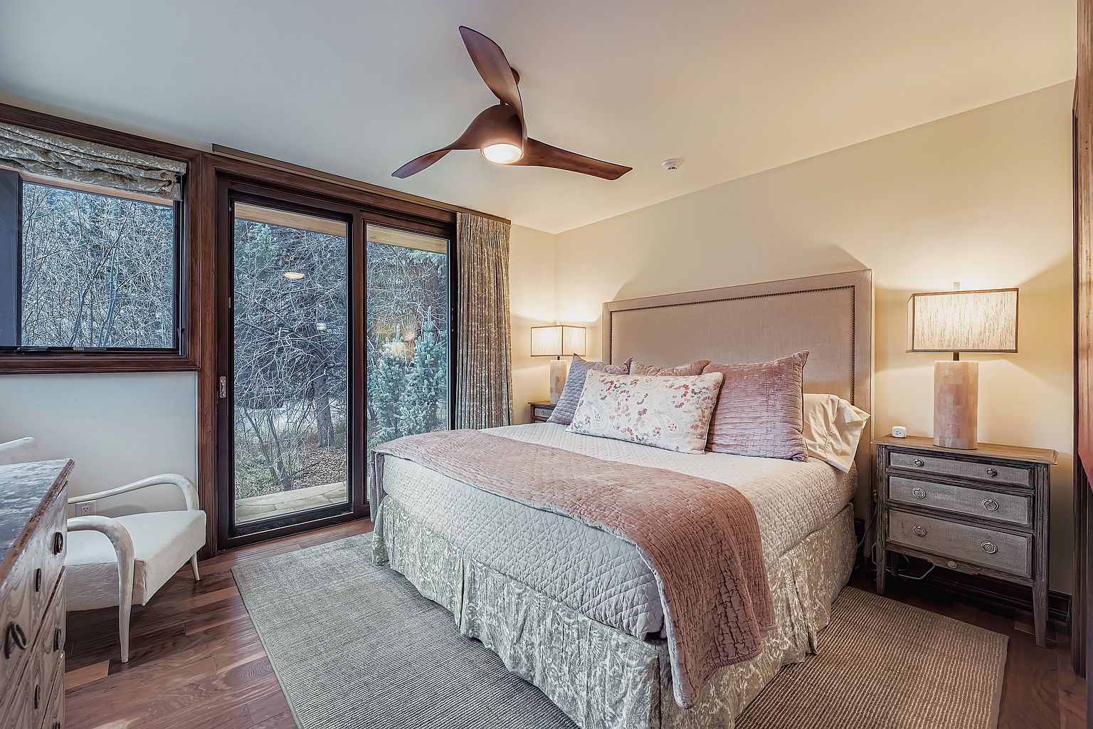 This is a cozy primary bedroom featuring a large bed with a padded headboard, decorative pillows, and a patterned bed skirt. The room has a ceiling fan, bedside tables with lamps, and windows offering a view of the outdoors. The overall style is comfortable and inviting, perfect for relaxation.