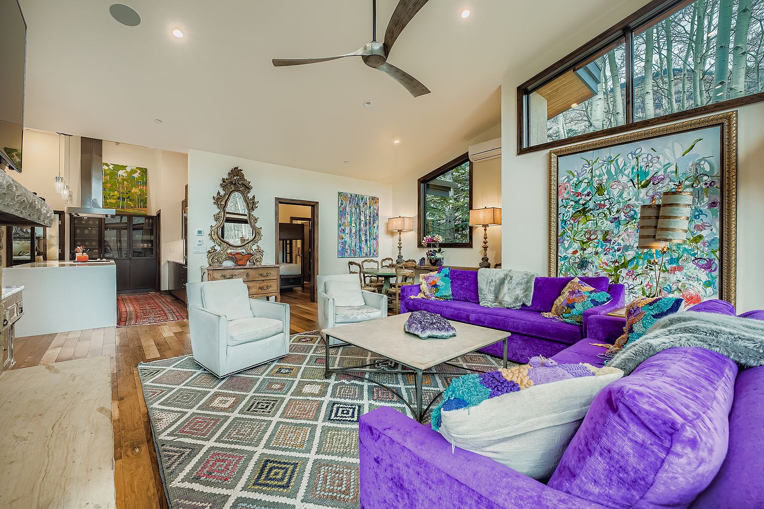 This is an interior shot of a living room featuring a vibrant purple sectional sofa, a patterned area rug, and two white armchairs. The room is decorated with eclectic artwork and a large ornate mirror, creating a luxurious and inviting atmosphere. Natural light streams in through the windows, highlighting the room's unique design elements.