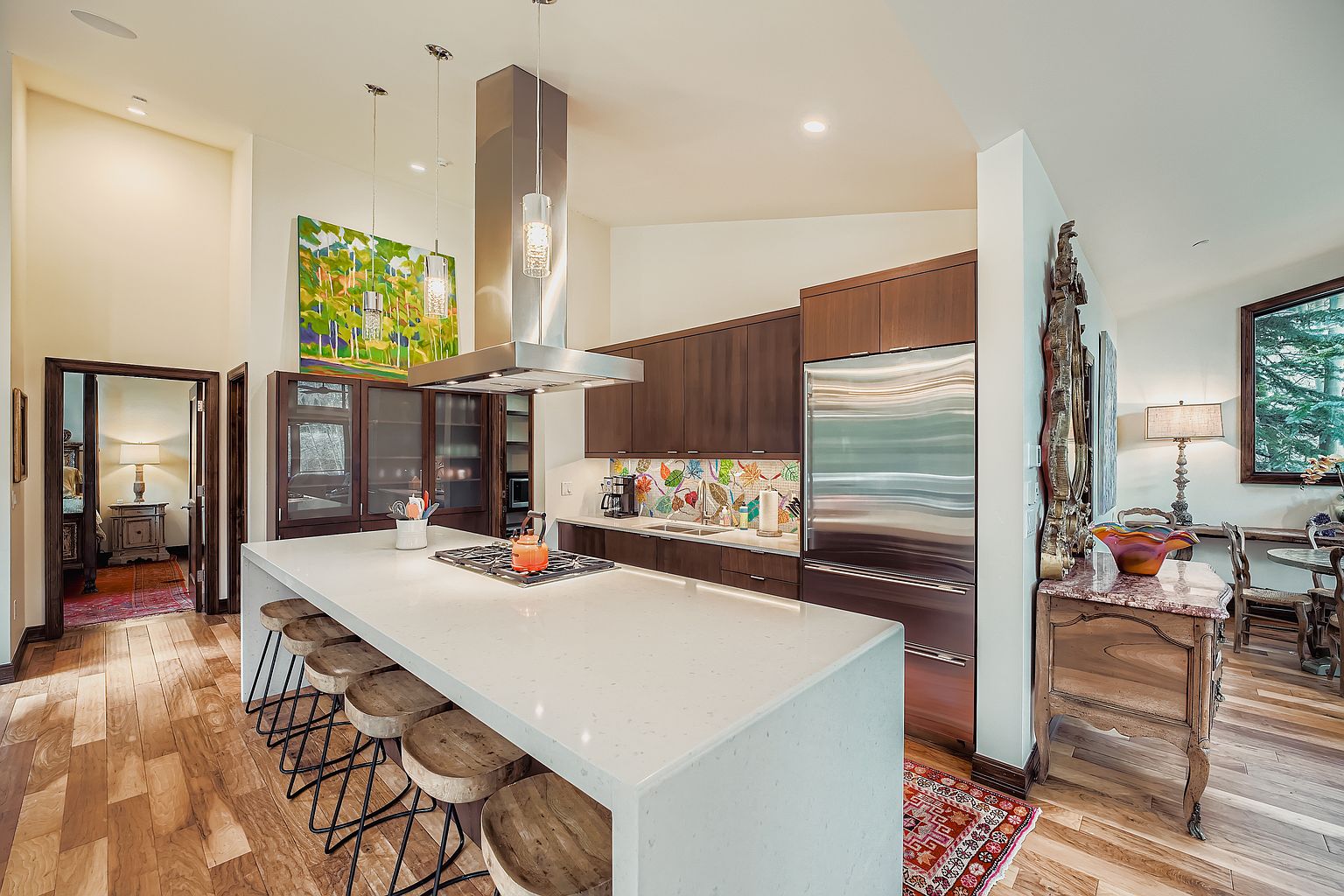 This is a well-lit kitchen featuring dark wood cabinetry, stainless steel appliances, and a large white countertop island with seating. The kitchen also has a colorful backsplash and a modern stainless steel range hood. The perspective is from a medium distance, showcasing the kitchen's layout and design elements.