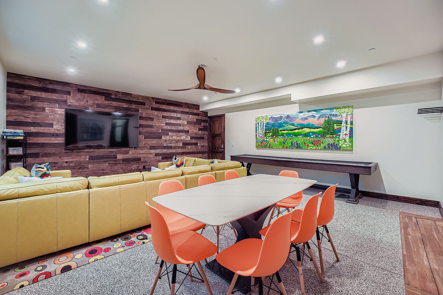 This interior shot showcases a dining area with a modern aesthetic. A large, rectangular dining table with a white marble top and a dark, sculptural base is surrounded by orange chairs. The room also features a yellow sectional sofa, a wood-paneled wall with a large television, and a colorful rug, creating a vibrant and inviting space.