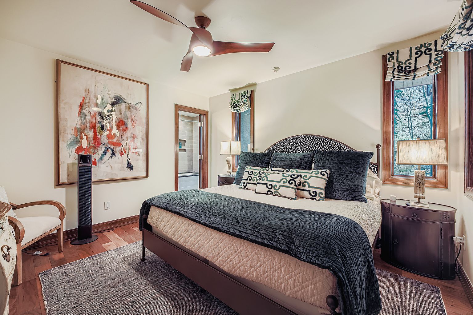 This is a primary bedroom featuring a large bed with a patterned headboard, several pillows, and a dark blue throw blanket. The room has hardwood floors, a ceiling fan, and an abstract painting on the wall. There is a window with patterned blinds and a nightstand with a lamp on one side of the bed. The perspective is from the foot of the bed, looking towards the headboard and the rest of the room.