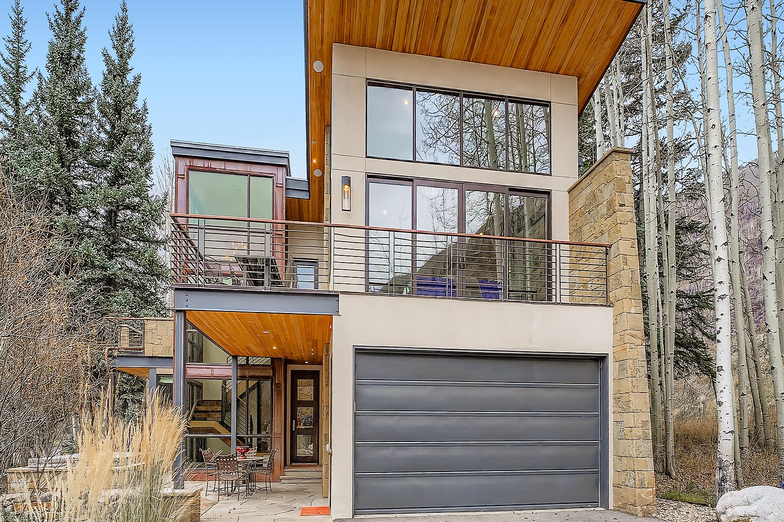 This is a front view of a modern, multi-level home with a contemporary design. The house features a combination of materials including wood, stone, and stucco, with large windows and balconies. A gray garage door is visible on the right side of the house, and landscaping elements such as tall grasses and trees surround the property, enhancing its curb appeal.