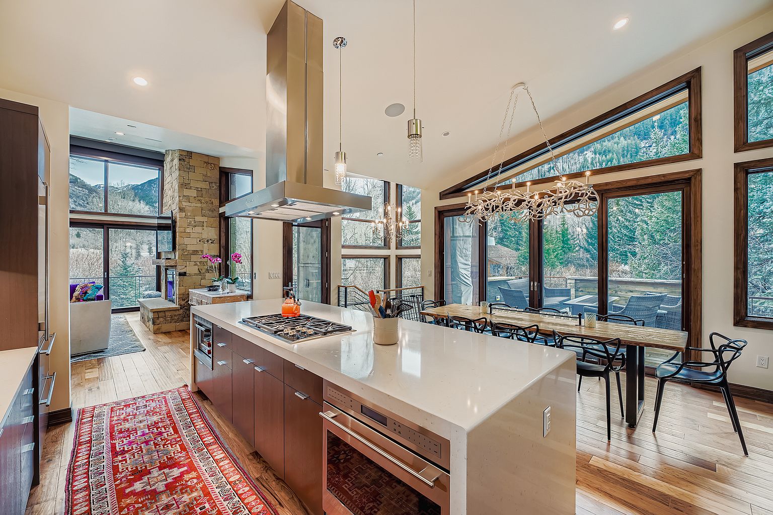 This is a bright and spacious kitchen featuring a large island with a cooktop and oven, modern cabinetry, and stainless steel appliances. The open floor plan connects to a dining area with a long wooden table and black chairs, all illuminated by a unique chandelier. Large windows offer stunning views of the surrounding landscape, creating a seamless indoor-outdoor living experience.