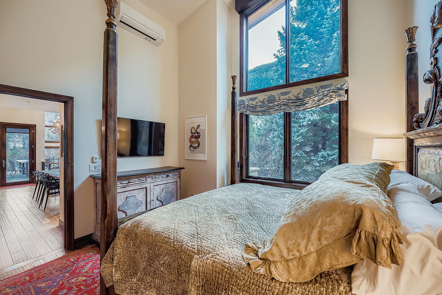 This is a primary bedroom featuring a four-poster bed with a decorative headboard and a neutral-toned quilt. A large window offers a view of the surrounding trees, and a console table with a television is positioned against the wall. The room has a cozy and luxurious feel, enhanced by the warm lighting and rich textures.