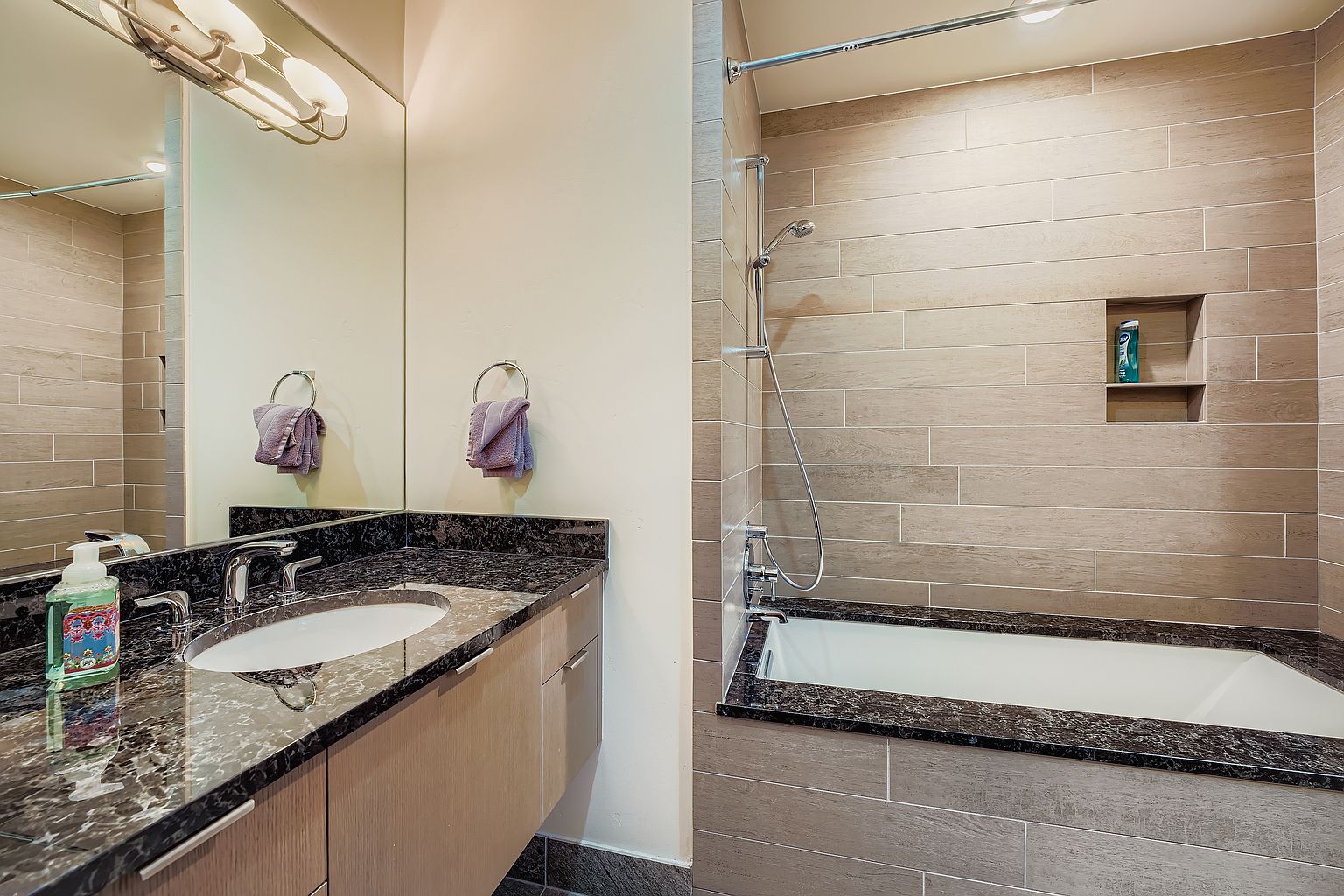 This is a bathroom featuring a vanity with a dark granite countertop and a light wood cabinet. A large mirror reflects the vanity and part of the tiled shower/tub area. The shower/tub is surrounded by horizontal tiles and has a built-in niche for toiletries, creating a modern and clean aesthetic.