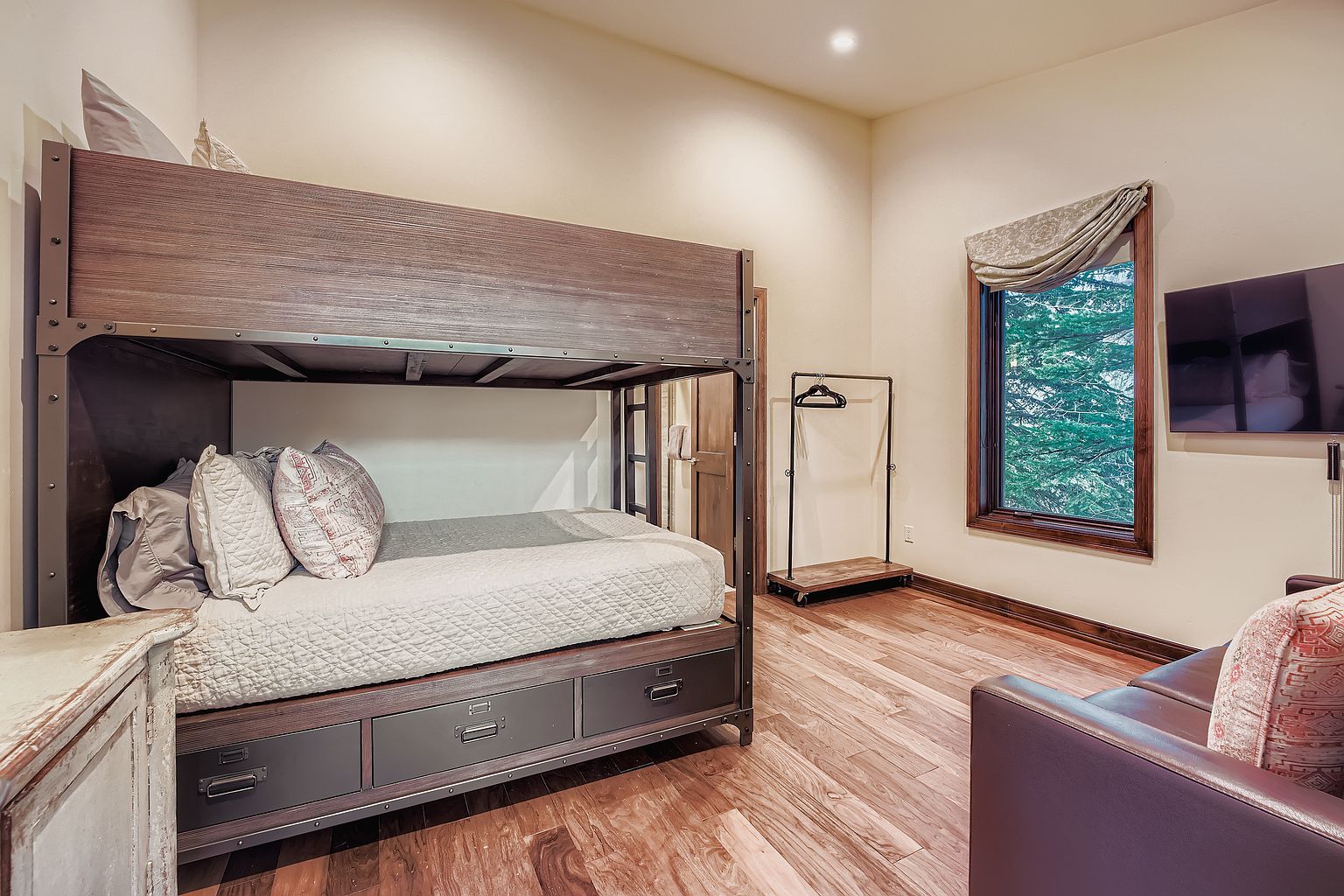This is a cozy bedroom featuring a unique bunk bed with a wooden frame and built-in storage drawers. The room has hardwood floors, a window with a decorative valance, and a television mounted on the wall. A clothing rack and a glimpse of a leather chair add to the room's functionality and comfort.
