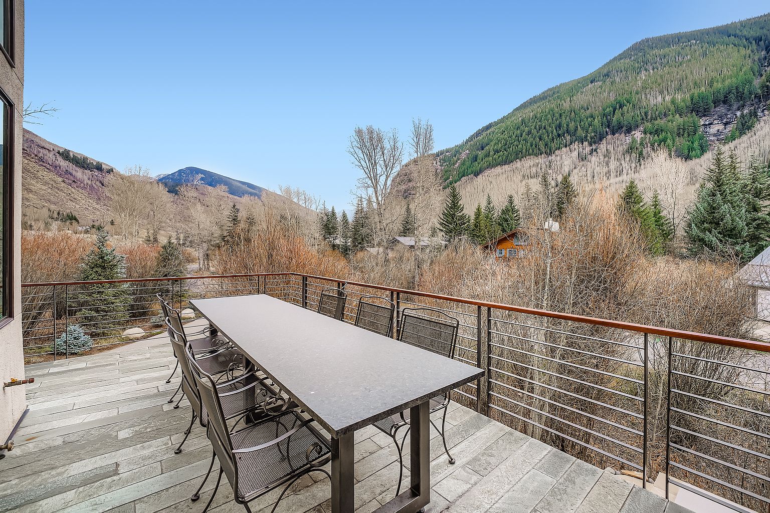 This image showcases a spacious outdoor patio or deck area, furnished with a large dining table and chairs, perfect for entertaining. The deck is surrounded by a railing, offering unobstructed views of the surrounding natural landscape, including mountains and trees. The setting suggests a tranquil and private outdoor living space.