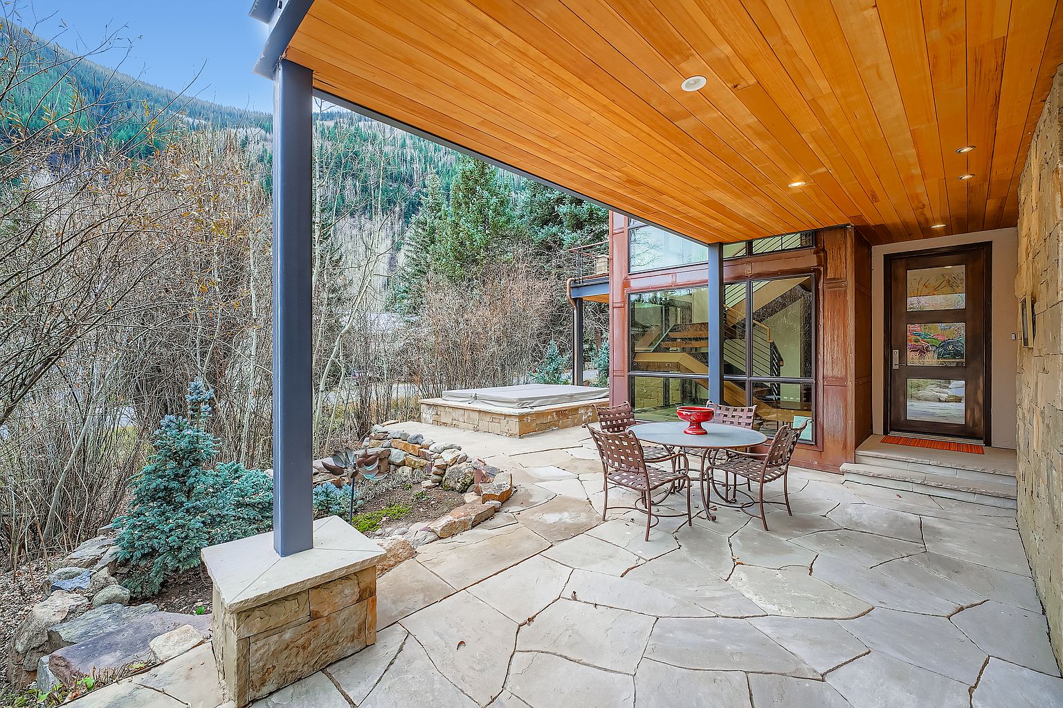 This image showcases a beautifully designed outdoor patio area. The space features a stone patio, a dining table with chairs, and a hot tub in the background. The wooden ceiling adds warmth, while the surrounding trees and mountain view create a serene and inviting atmosphere, perfect for relaxation and entertaining.
