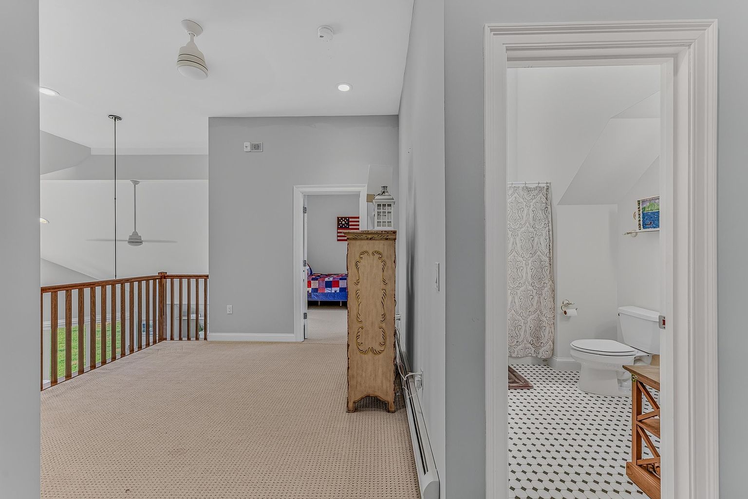 This image captures a carpeted landing area that overlooks a lower level, featuring a wooden railing and a ceiling fan. To the right, a doorway leads into a bathroom with patterned tile flooring and a shower curtain, while a straight path ahead leads to a bedroom visible in the distance. A tall, decorative wooden cabinet stands against the wall, adding a traditional touch to the neutral-toned space.