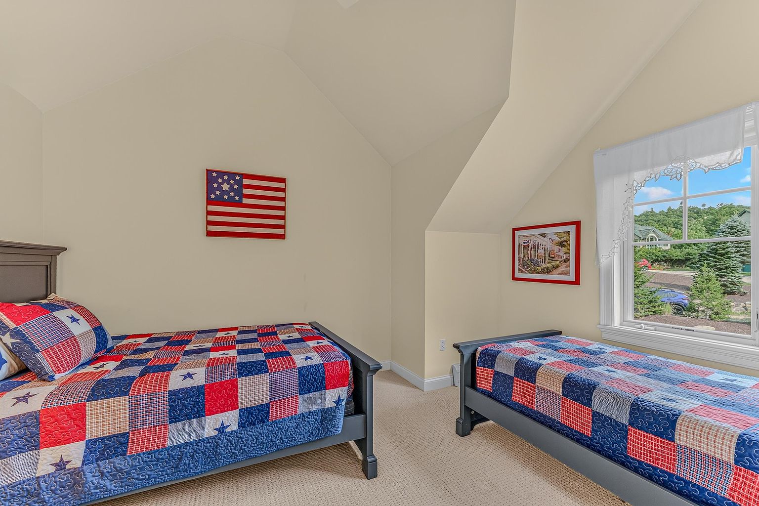 This guest bedroom features two twin beds with matching patriotic-themed patchwork quilts and pillows. The room has light-colored walls, a vaulted ceiling, and a large window that provides natural light and a view of the outdoors. A framed picture hangs on the wall, and a wooden American flag wall art piece adds to the Americana decor theme.