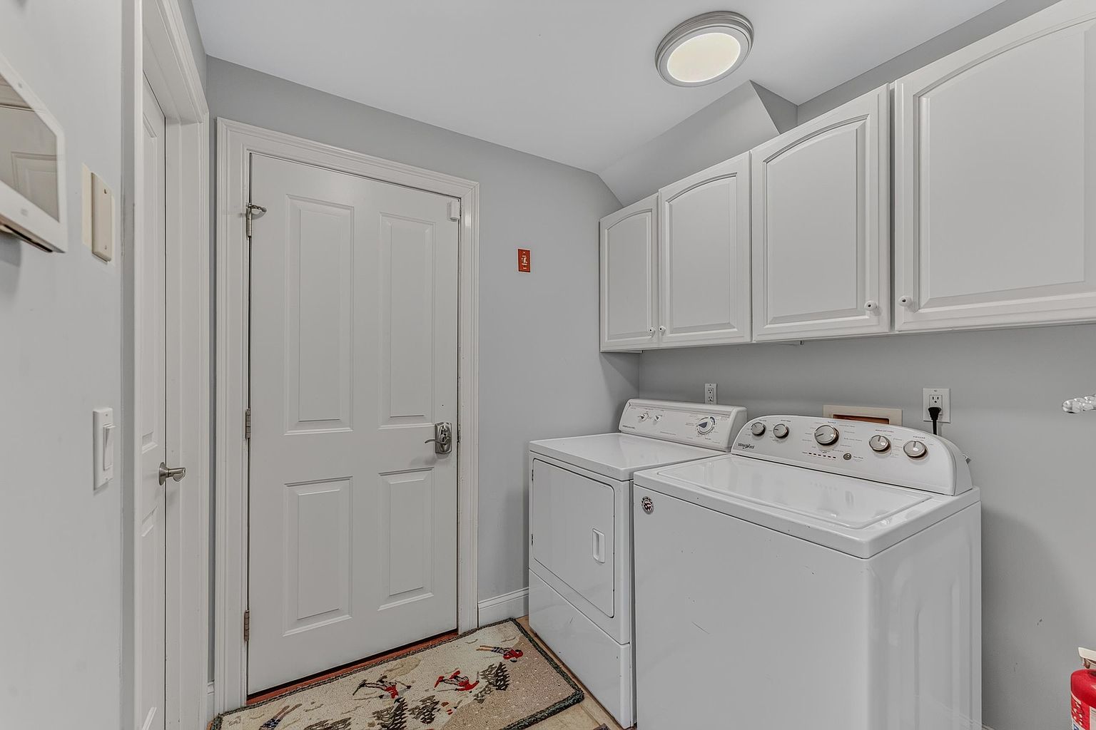 This functional laundry room features a side-by-side washer and dryer set positioned beneath white upper cabinetry that provides ample storage. A white door leads to the exterior, and a patterned rug adds a touch of character to the floor space. The room is illuminated by a simple flush-mount ceiling light, creating a clean and practical utility area.