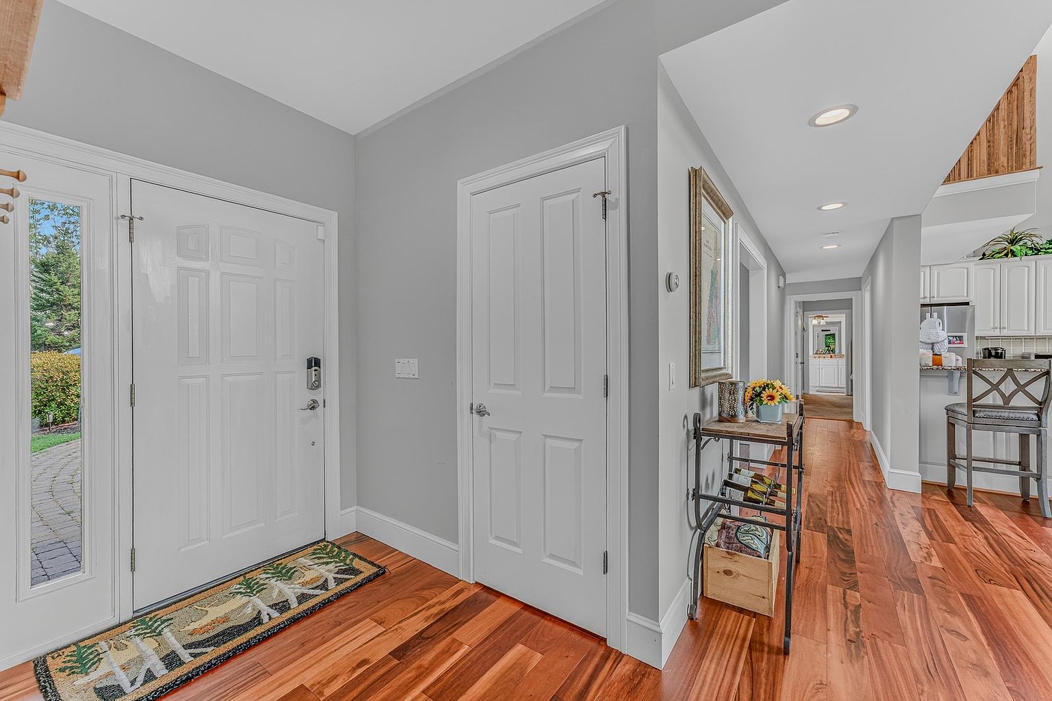 This bright and welcoming entryway features rich, warm-toned wood flooring that extends into a long hallway. The space includes a white front door with glass sidelights, a neutral gray wall color, and a decorative metal console table holding a small floral arrangement. The perspective looks from the front door down the hallway, offering a clear view into the adjacent kitchen area.