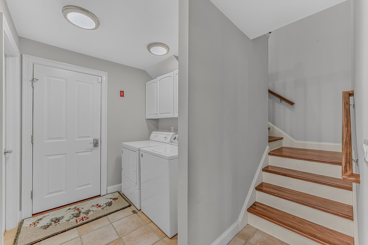 This interior view captures a functional laundry area situated adjacent to a staircase. The space features a white washer and dryer set tucked beneath wall-mounted cabinets, with a light-colored tile floor and a ski-themed rug near the door. To the right, a wooden staircase with a handrail leads upward, creating a practical transition space within the home.
